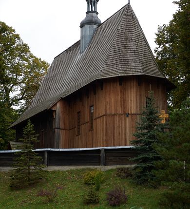 Zdjęcie nr 6: Kościół drewniany, orientowany, usytuowany na wzgórku, otoczony pierwotnym cmentarzem przykościelnym oraz starodrzewiem (lipami), ujęty drewnianym ogrodzeniem z poziomo ułożonych desek, w którego ciągu znajduje się masywna dzwonnica. Ściany kościoła wzniesione w konstrukcji zrębowej, oszalowanej pionowo z listwowaniem. Nawa na planie prostokąta; prezbiterium węższe, zamknięte wielobocznie, zbudowane na zrąb; od północy zakrystia na planie prostokąta; od południa późniejsza, dobudowana kruchta. Od zachodu nawę poprzedza niższa wieża, zbudowana w konstrukcji słupowo-ramowej, o pochyłych ścianach. Wnętrze nakryte stropami, w nawie z zaskrzynieniami. Nawa od prezbiterium oddzielona tęczą o łuku półkolistym wspartym na kroksztynach; na belce umieszczone rzeźby z początku XVI wieku przedstawiające grupę Ukrzyżowania: krucyfiks, Matka Boska, św. Jan Ewangelista oraz św. Maria Magdalena. Chór muzyczny wsparty na dwóch kolumnach, barokowy. Wnętrze pokryte polichromią z pierwszej połowy XVII wieku. Pośrodku kościoła oraz bliżej chóru muzycznego znajdują się wejścia do krypt pokryte płytami. Główny portal kościoła umieszczony jest od strony zachodniej, w przedsionku pod wieżą, ukształtowany w formie profilowanej, lekko zaostrzonej arkady przechodzącej w tzw. ośli grzbiet. Portal południowy, w kruchcie zakończony ostrołukiem, z motywem sznura w profilowaniach z pierwotnymi jednoskrzydłowymi drzwiami o gotyckich okuciach. Portal prowadzący z prezbiterium do zakrystii ma bogate profilowanie (zewnętrzny profil z motywem „ząbków”), jest zakończony w tzw. ośli grzbiet z drzwiami żelaznymi, dekorowanymi ukośną kratą. Okna znajdują się od strony południowej, pod dwa w nawie oraz w prezbiterium. Nadokienniki mają wykrój w ośli grzbiet. Ponadto jedno okno o analogicznym układzie w prezbiterium na ścianie wschodniej, częściowo zabudowane, zasłonięte ołtarzem. Dach gontowy o jednej kalenicy, wsparty na kroksztynach z szerokim okapem, o dużym kącie spadku uzyskanym poprzez zastosowaną gotycką więźbę storczykową, pośrodku z wieżyczką na sygnaturkę o barkowym kształcie. Wieża główna nakryta dachem gontowym, trójspadowym.
