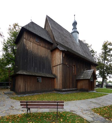 Zdjęcie nr 10: Kościół drewniany, orientowany, usytuowany na wzgórku, otoczony pierwotnym cmentarzem przykościelnym oraz starodrzewiem (lipami), ujęty drewnianym ogrodzeniem z poziomo ułożonych desek, w którego ciągu znajduje się masywna dzwonnica. Ściany kościoła wzniesione w konstrukcji zrębowej, oszalowanej pionowo z listwowaniem. Nawa na planie prostokąta; prezbiterium węższe, zamknięte wielobocznie, zbudowane na zrąb; od północy zakrystia na planie prostokąta; od południa późniejsza, dobudowana kruchta. Od zachodu nawę poprzedza niższa wieża, zbudowana w konstrukcji słupowo-ramowej, o pochyłych ścianach. Wnętrze nakryte stropami, w nawie z zaskrzynieniami. Nawa od prezbiterium oddzielona tęczą o łuku półkolistym wspartym na kroksztynach; na belce umieszczone rzeźby z początku XVI wieku przedstawiające grupę Ukrzyżowania: krucyfiks, Matka Boska, św. Jan Ewangelista oraz św. Maria Magdalena. Chór muzyczny wsparty na dwóch kolumnach, barokowy. Wnętrze pokryte polichromią z pierwszej połowy XVII wieku. Pośrodku kościoła oraz bliżej chóru muzycznego znajdują się wejścia do krypt pokryte płytami. Główny portal kościoła umieszczony jest od strony zachodniej, w przedsionku pod wieżą, ukształtowany w formie profilowanej, lekko zaostrzonej arkady przechodzącej w tzw. ośli grzbiet. Portal południowy, w kruchcie zakończony ostrołukiem, z motywem sznura w profilowaniach z pierwotnymi jednoskrzydłowymi drzwiami o gotyckich okuciach. Portal prowadzący z prezbiterium do zakrystii ma bogate profilowanie (zewnętrzny profil z motywem „ząbków”), jest zakończony w tzw. ośli grzbiet z drzwiami żelaznymi, dekorowanymi ukośną kratą. Okna znajdują się od strony południowej, pod dwa w nawie oraz w prezbiterium. Nadokienniki mają wykrój w ośli grzbiet. Ponadto jedno okno o analogicznym układzie w prezbiterium na ścianie wschodniej, częściowo zabudowane, zasłonięte ołtarzem. Dach gontowy o jednej kalenicy, wsparty na kroksztynach z szerokim okapem, o dużym kącie spadku uzyskanym poprzez zastosowaną gotycką więźbę storczykową, pośrodku z wieżyczką na sygnaturkę o barkowym kształcie. Wieża główna nakryta dachem gontowym, trójspadowym.
