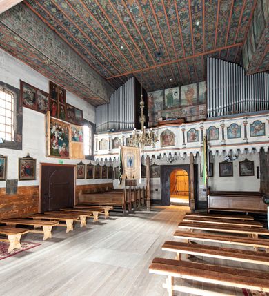 Zdjęcie nr 19: Kościół drewniany, orientowany, usytuowany na wzgórku, otoczony pierwotnym cmentarzem przykościelnym oraz starodrzewiem (lipami), ujęty drewnianym ogrodzeniem z poziomo ułożonych desek, w którego ciągu znajduje się masywna dzwonnica. Ściany kościoła wzniesione w konstrukcji zrębowej, oszalowanej pionowo z listwowaniem. Nawa na planie prostokąta; prezbiterium węższe, zamknięte wielobocznie, zbudowane na zrąb; od północy zakrystia na planie prostokąta; od południa późniejsza, dobudowana kruchta. Od zachodu nawę poprzedza niższa wieża, zbudowana w konstrukcji słupowo-ramowej, o pochyłych ścianach. Wnętrze nakryte stropami, w nawie z zaskrzynieniami. Nawa od prezbiterium oddzielona tęczą o łuku półkolistym wspartym na kroksztynach; na belce umieszczone rzeźby z początku XVI wieku przedstawiające grupę Ukrzyżowania: krucyfiks, Matka Boska, św. Jan Ewangelista oraz św. Maria Magdalena. Chór muzyczny wsparty na dwóch kolumnach, barokowy. Wnętrze pokryte polichromią z pierwszej połowy XVII wieku. Pośrodku kościoła oraz bliżej chóru muzycznego znajdują się wejścia do krypt pokryte płytami. Główny portal kościoła umieszczony jest od strony zachodniej, w przedsionku pod wieżą, ukształtowany w formie profilowanej, lekko zaostrzonej arkady przechodzącej w tzw. ośli grzbiet. Portal południowy, w kruchcie zakończony ostrołukiem, z motywem sznura w profilowaniach z pierwotnymi jednoskrzydłowymi drzwiami o gotyckich okuciach. Portal prowadzący z prezbiterium do zakrystii ma bogate profilowanie (zewnętrzny profil z motywem „ząbków”), jest zakończony w tzw. ośli grzbiet z drzwiami żelaznymi, dekorowanymi ukośną kratą. Okna znajdują się od strony południowej, pod dwa w nawie oraz w prezbiterium. Nadokienniki mają wykrój w ośli grzbiet. Ponadto jedno okno o analogicznym układzie w prezbiterium na ścianie wschodniej, częściowo zabudowane, zasłonięte ołtarzem. Dach gontowy o jednej kalenicy, wsparty na kroksztynach z szerokim okapem, o dużym kącie spadku uzyskanym poprzez zastosowaną gotycką więźbę storczykową, pośrodku z wieżyczką na sygnaturkę o barkowym kształcie. Wieża główna nakryta dachem gontowym, trójspadowym.
