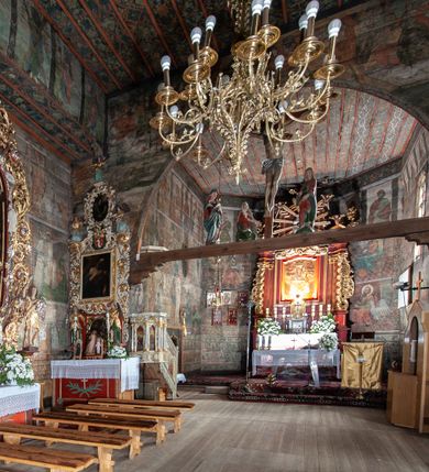 Zdjęcie nr 13: Kościół drewniany, orientowany, usytuowany na wzgórku, otoczony pierwotnym cmentarzem przykościelnym oraz starodrzewiem (lipami), ujęty drewnianym ogrodzeniem z poziomo ułożonych desek, w którego ciągu znajduje się masywna dzwonnica. Ściany kościoła wzniesione w konstrukcji zrębowej, oszalowanej pionowo z listwowaniem. Nawa na planie prostokąta; prezbiterium węższe, zamknięte wielobocznie, zbudowane na zrąb; od północy zakrystia na planie prostokąta; od południa późniejsza, dobudowana kruchta. Od zachodu nawę poprzedza niższa wieża, zbudowana w konstrukcji słupowo-ramowej, o pochyłych ścianach. Wnętrze nakryte stropami, w nawie z zaskrzynieniami. Nawa od prezbiterium oddzielona tęczą o łuku półkolistym wspartym na kroksztynach; na belce umieszczone rzeźby z początku XVI wieku przedstawiające grupę Ukrzyżowania: krucyfiks, Matka Boska, św. Jan Ewangelista oraz św. Maria Magdalena. Chór muzyczny wsparty na dwóch kolumnach, barokowy. Wnętrze pokryte polichromią z pierwszej połowy XVII wieku. Pośrodku kościoła oraz bliżej chóru muzycznego znajdują się wejścia do krypt pokryte płytami. Główny portal kościoła umieszczony jest od strony zachodniej, w przedsionku pod wieżą, ukształtowany w formie profilowanej, lekko zaostrzonej arkady przechodzącej w tzw. ośli grzbiet. Portal południowy, w kruchcie zakończony ostrołukiem, z motywem sznura w profilowaniach z pierwotnymi jednoskrzydłowymi drzwiami o gotyckich okuciach. Portal prowadzący z prezbiterium do zakrystii ma bogate profilowanie (zewnętrzny profil z motywem „ząbków”), jest zakończony w tzw. ośli grzbiet z drzwiami żelaznymi, dekorowanymi ukośną kratą. Okna znajdują się od strony południowej, pod dwa w nawie oraz w prezbiterium. Nadokienniki mają wykrój w ośli grzbiet. Ponadto jedno okno o analogicznym układzie w prezbiterium na ścianie wschodniej, częściowo zabudowane, zasłonięte ołtarzem. Dach gontowy o jednej kalenicy, wsparty na kroksztynach z szerokim okapem, o dużym kącie spadku uzyskanym poprzez zastosowaną gotycką więźbę storczykową, pośrodku z wieżyczką na sygnaturkę o barkowym kształcie. Wieża główna nakryta dachem gontowym, trójspadowym.
