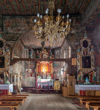 Zdjęcie nr 12: Kościół drewniany, orientowany, usytuowany na wzgórku, otoczony pierwotnym cmentarzem przykościelnym oraz starodrzewiem (lipami), ujęty drewnianym ogrodzeniem z poziomo ułożonych desek, w którego ciągu znajduje się masywna dzwonnica. Ściany kościoła wzniesione w konstrukcji zrębowej, oszalowanej pionowo z listwowaniem. Nawa na planie prostokąta; prezbiterium węższe, zamknięte wielobocznie, zbudowane na zrąb; od północy zakrystia na planie prostokąta; od południa późniejsza, dobudowana kruchta. Od zachodu nawę poprzedza niższa wieża, zbudowana w konstrukcji słupowo-ramowej, o pochyłych ścianach. Wnętrze nakryte stropami, w nawie z zaskrzynieniami. Nawa od prezbiterium oddzielona tęczą o łuku półkolistym wspartym na kroksztynach; na belce umieszczone rzeźby z początku XVI wieku przedstawiające grupę Ukrzyżowania: krucyfiks, Matka Boska, św. Jan Ewangelista oraz św. Maria Magdalena. Chór muzyczny wsparty na dwóch kolumnach, barokowy. Wnętrze pokryte polichromią z pierwszej połowy XVII wieku. Pośrodku kościoła oraz bliżej chóru muzycznego znajdują się wejścia do krypt pokryte płytami. Główny portal kościoła umieszczony jest od strony zachodniej, w przedsionku pod wieżą, ukształtowany w formie profilowanej, lekko zaostrzonej arkady przechodzącej w tzw. ośli grzbiet. Portal południowy, w kruchcie zakończony ostrołukiem, z motywem sznura w profilowaniach z pierwotnymi jednoskrzydłowymi drzwiami o gotyckich okuciach. Portal prowadzący z prezbiterium do zakrystii ma bogate profilowanie (zewnętrzny profil z motywem „ząbków”), jest zakończony w tzw. ośli grzbiet z drzwiami żelaznymi, dekorowanymi ukośną kratą. Okna znajdują się od strony południowej, pod dwa w nawie oraz w prezbiterium. Nadokienniki mają wykrój w ośli grzbiet. Ponadto jedno okno o analogicznym układzie w prezbiterium na ścianie wschodniej, częściowo zabudowane, zasłonięte ołtarzem. Dach gontowy o jednej kalenicy, wsparty na kroksztynach z szerokim okapem, o dużym kącie spadku uzyskanym poprzez zastosowaną gotycką więźbę storczykową, pośrodku z wieżyczką na sygnaturkę o barkowym kształcie. Wieża główna nakryta dachem gontowym, trójspadowym.
