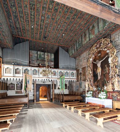 Zdjęcie nr 18: Kościół drewniany, orientowany, usytuowany na wzgórku, otoczony pierwotnym cmentarzem przykościelnym oraz starodrzewiem (lipami), ujęty drewnianym ogrodzeniem z poziomo ułożonych desek, w którego ciągu znajduje się masywna dzwonnica. Ściany kościoła wzniesione w konstrukcji zrębowej, oszalowanej pionowo z listwowaniem. Nawa na planie prostokąta; prezbiterium węższe, zamknięte wielobocznie, zbudowane na zrąb; od północy zakrystia na planie prostokąta; od południa późniejsza, dobudowana kruchta. Od zachodu nawę poprzedza niższa wieża, zbudowana w konstrukcji słupowo-ramowej, o pochyłych ścianach. Wnętrze nakryte stropami, w nawie z zaskrzynieniami. Nawa od prezbiterium oddzielona tęczą o łuku półkolistym wspartym na kroksztynach; na belce umieszczone rzeźby z początku XVI wieku przedstawiające grupę Ukrzyżowania: krucyfiks, Matka Boska, św. Jan Ewangelista oraz św. Maria Magdalena. Chór muzyczny wsparty na dwóch kolumnach, barokowy. Wnętrze pokryte polichromią z pierwszej połowy XVII wieku. Pośrodku kościoła oraz bliżej chóru muzycznego znajdują się wejścia do krypt pokryte płytami. Główny portal kościoła umieszczony jest od strony zachodniej, w przedsionku pod wieżą, ukształtowany w formie profilowanej, lekko zaostrzonej arkady przechodzącej w tzw. ośli grzbiet. Portal południowy, w kruchcie zakończony ostrołukiem, z motywem sznura w profilowaniach z pierwotnymi jednoskrzydłowymi drzwiami o gotyckich okuciach. Portal prowadzący z prezbiterium do zakrystii ma bogate profilowanie (zewnętrzny profil z motywem „ząbków”), jest zakończony w tzw. ośli grzbiet z drzwiami żelaznymi, dekorowanymi ukośną kratą. Okna znajdują się od strony południowej, pod dwa w nawie oraz w prezbiterium. Nadokienniki mają wykrój w ośli grzbiet. Ponadto jedno okno o analogicznym układzie w prezbiterium na ścianie wschodniej, częściowo zabudowane, zasłonięte ołtarzem. Dach gontowy o jednej kalenicy, wsparty na kroksztynach z szerokim okapem, o dużym kącie spadku uzyskanym poprzez zastosowaną gotycką więźbę storczykową, pośrodku z wieżyczką na sygnaturkę o barkowym kształcie. Wieża główna nakryta dachem gontowym, trójspadowym.
