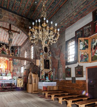 Zdjęcie nr 14: Kościół drewniany, orientowany, usytuowany na wzgórku, otoczony pierwotnym cmentarzem przykościelnym oraz starodrzewiem (lipami), ujęty drewnianym ogrodzeniem z poziomo ułożonych desek, w którego ciągu znajduje się masywna dzwonnica. Ściany kościoła wzniesione w konstrukcji zrębowej, oszalowanej pionowo z listwowaniem. Nawa na planie prostokąta; prezbiterium węższe, zamknięte wielobocznie, zbudowane na zrąb; od północy zakrystia na planie prostokąta; od południa późniejsza, dobudowana kruchta. Od zachodu nawę poprzedza niższa wieża, zbudowana w konstrukcji słupowo-ramowej, o pochyłych ścianach. Wnętrze nakryte stropami, w nawie z zaskrzynieniami. Nawa od prezbiterium oddzielona tęczą o łuku półkolistym wspartym na kroksztynach; na belce umieszczone rzeźby z początku XVI wieku przedstawiające grupę Ukrzyżowania: krucyfiks, Matka Boska, św. Jan Ewangelista oraz św. Maria Magdalena. Chór muzyczny wsparty na dwóch kolumnach, barokowy. Wnętrze pokryte polichromią z pierwszej połowy XVII wieku. Pośrodku kościoła oraz bliżej chóru muzycznego znajdują się wejścia do krypt pokryte płytami. Główny portal kościoła umieszczony jest od strony zachodniej, w przedsionku pod wieżą, ukształtowany w formie profilowanej, lekko zaostrzonej arkady przechodzącej w tzw. ośli grzbiet. Portal południowy, w kruchcie zakończony ostrołukiem, z motywem sznura w profilowaniach z pierwotnymi jednoskrzydłowymi drzwiami o gotyckich okuciach. Portal prowadzący z prezbiterium do zakrystii ma bogate profilowanie (zewnętrzny profil z motywem „ząbków”), jest zakończony w tzw. ośli grzbiet z drzwiami żelaznymi, dekorowanymi ukośną kratą. Okna znajdują się od strony południowej, pod dwa w nawie oraz w prezbiterium. Nadokienniki mają wykrój w ośli grzbiet. Ponadto jedno okno o analogicznym układzie w prezbiterium na ścianie wschodniej, częściowo zabudowane, zasłonięte ołtarzem. Dach gontowy o jednej kalenicy, wsparty na kroksztynach z szerokim okapem, o dużym kącie spadku uzyskanym poprzez zastosowaną gotycką więźbę storczykową, pośrodku z wieżyczką na sygnaturkę o barkowym kształcie. Wieża główna nakryta dachem gontowym, trójspadowym.
