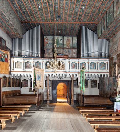 Zdjęcie nr 17: Kościół drewniany, orientowany, usytuowany na wzgórku, otoczony pierwotnym cmentarzem przykościelnym oraz starodrzewiem (lipami), ujęty drewnianym ogrodzeniem z poziomo ułożonych desek, w którego ciągu znajduje się masywna dzwonnica. Ściany kościoła wzniesione w konstrukcji zrębowej, oszalowanej pionowo z listwowaniem. Nawa na planie prostokąta; prezbiterium węższe, zamknięte wielobocznie, zbudowane na zrąb; od północy zakrystia na planie prostokąta; od południa późniejsza, dobudowana kruchta. Od zachodu nawę poprzedza niższa wieża, zbudowana w konstrukcji słupowo-ramowej, o pochyłych ścianach. Wnętrze nakryte stropami, w nawie z zaskrzynieniami. Nawa od prezbiterium oddzielona tęczą o łuku półkolistym wspartym na kroksztynach; na belce umieszczone rzeźby z początku XVI wieku przedstawiające grupę Ukrzyżowania: krucyfiks, Matka Boska, św. Jan Ewangelista oraz św. Maria Magdalena. Chór muzyczny wsparty na dwóch kolumnach, barokowy. Wnętrze pokryte polichromią z pierwszej połowy XVII wieku. Pośrodku kościoła oraz bliżej chóru muzycznego znajdują się wejścia do krypt pokryte płytami. Główny portal kościoła umieszczony jest od strony zachodniej, w przedsionku pod wieżą, ukształtowany w formie profilowanej, lekko zaostrzonej arkady przechodzącej w tzw. ośli grzbiet. Portal południowy, w kruchcie zakończony ostrołukiem, z motywem sznura w profilowaniach z pierwotnymi jednoskrzydłowymi drzwiami o gotyckich okuciach. Portal prowadzący z prezbiterium do zakrystii ma bogate profilowanie (zewnętrzny profil z motywem „ząbków”), jest zakończony w tzw. ośli grzbiet z drzwiami żelaznymi, dekorowanymi ukośną kratą. Okna znajdują się od strony południowej, pod dwa w nawie oraz w prezbiterium. Nadokienniki mają wykrój w ośli grzbiet. Ponadto jedno okno o analogicznym układzie w prezbiterium na ścianie wschodniej, częściowo zabudowane, zasłonięte ołtarzem. Dach gontowy o jednej kalenicy, wsparty na kroksztynach z szerokim okapem, o dużym kącie spadku uzyskanym poprzez zastosowaną gotycką więźbę storczykową, pośrodku z wieżyczką na sygnaturkę o barkowym kształcie. Wieża główna nakryta dachem gontowym, trójspadowym.
