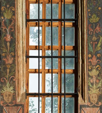 Zdjęcie nr 2: Polichromia ścienna wokół okien prostokątnych, zamkniętych w tzw. ośli grzbiet tworzy kompozycje o kształcie stojących prostokątów. Po bokach obramienia w dolnej części znajdują się stojące prostokąty przecięte liniami na kształt litery „X”. Powyżej umieszczone są maski o ludzkich twarzach z wolutowo zawiniętymi uszami i liśćmi akantu zamiast włosów na czubku głowy. W miejscu szyi mają dwie czerwone wolutki. Powyżej w układzie kandelabrowym wyrastają liście, motywy kampanul i kółka. Liście akantu zdobią również górne naroża obramowań. Całość ukazana jest na czarnym tle. Motywy dekoracyjne o intensywnych barwach, różnokolorowych z przewagą zieleni i czerwieni, obwiedzione grubym i czarnym konturem. 