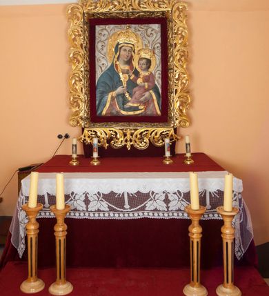 Zdjęcie nr 10: Obraz w formie stojącego prostokąta. Maria ukazana w półpostaci, zwrócona w trzech czwartych w lewo. W prawej, ugiętej ręce trzyma berło, lewą podtrzymuje Dzieciątko. Twarz ma owalną, o łagodnych rysach, migdałowatych oczach, długim nosie i pełnych ustach, okoloną jasnobrązowymi włosami. Ubrana jest w jasnoczerwoną suknię ze złoconymi lamówkami oraz granatowy maforion z czerwoną podszewką i złoconą lamówką zarzucony na głowę i ramiona, zapięty na piersi okrągłą klamrą. Dzieciątko ukazane w pozycji siedzącej, skierowane w trzech czwartych w prawo, z wysuniętą lewą nogą. Prawą rękę unosi w geście błogosławieństwa, lewą trzyma jabłko królewskie. Twarz ma kwadratową, o pełnych policzkach, dużych, migdałowatych oczach, małym nosie i pełnych ustach, okoloną krótkimi, brązowymi włosami. Ubrane jest w jasnoczerwoną tunikę ze złoconymi lamówkami, na stopach ma sandały. Maria i Dzieciątko mają na głowach zamknięte korony, a wokół nich nimby. Postacie ukazane są na srebrzonym tle wypełnionym wicią roślinną. Korony i berło rzeźbione są w drewnie, nałożone na płótno.