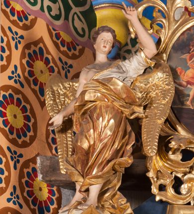 Zdjęcie nr 1: Figura anioła ukazana jest frontalnie, w kontrapoście, z uniesioną wysoko lewą ręką i dłonią w geście wskazującym oraz prawą ręką wysuniętą w bok. Twarz ma pociągłą, o wysokim czole, małych, migdałowatych oczach, nosie i otwartych ustach, okoloną krótkimi, falowanymi włosami. Anioł ubrany jest w białą tunikę ze złoconą lamówką, odsłaniającą połowę klatki piersiowej i prawe ramię oraz złocony, obfity płaszcz otaczający postać, ekspresyjnie drapowany, opadający na ziemię. Skrzydła ma złocone, złożone po bokach postaci. Polichromia ciała naturalistyczna. 