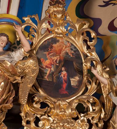 Zdjęcie nr 2: Figura anioła ukazana jest frontalnie, w kontrapoście, z uniesioną wysoko lewą ręką i dłonią w geście wskazującym oraz prawą ręką wysuniętą w bok. Twarz ma pociągłą, o wysokim czole, małych, migdałowatych oczach, nosie i otwartych ustach, okoloną krótkimi, falowanymi włosami. Anioł ubrany jest w białą tunikę ze złoconą lamówką, odsłaniającą połowę klatki piersiowej i prawe ramię oraz złocony, obfity płaszcz otaczający postać, ekspresyjnie drapowany, opadający na ziemię. Skrzydła ma złocone, złożone po bokach postaci. Polichromia ciała naturalistyczna. 