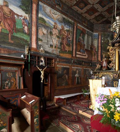 Zdjęcie nr 41: Polichromia ścienna prezbiterium pokrywa wszystkie ściany oraz strop tej części świątyni. 

Polichromia ścienna stropu
Strop zdobiony jest malowanymi kasetonami, w których umieszczone są rozety na różnokolorowych tłach. W centrum ukazane są dwie prostokątne kompozycje, ujęte w ornamentalne ramy. Od wschodu znajduje się Zwiastowanie Zachariaszowi, powyżej inskrypcja: „NE TIMEAS ZACHARIA VXOR TVA PARIET TIBI / FILIVM ET VOCABIS NOMEN EIVS IOANNEM LUCAE 1”. W drugiej kompozycji przedstawiono Nawiedzenie z Najświętszej Marii Panny ze św. Zachariaszem i św. Józefem, powyżej inskrypcja: „EXVLTAVIT IN GAVDIO INFANS IN VTERO MEO LUCAE 1”. 

Polichromia ścienna ścian prezbiterium
Dekoracje ścian o formie pasowej. W dolnej kondygnacji malowane iluzjonistycznie portrety świętych węgierskich w ornamentalnych ramach. Postacie ukazane do wysokości kolan, zwrócone trzy czwarte z odpowiednimi im atrybutami w dłoniach. Portrety opatrzone u góry łacińskimi opisami. Na ścianie północnej galerię otwiera wizerunek króla Stefana z napisem: „S(ANCTVS) STEPHANVS REX VNGARIAE, REGNVM SVVM DEIPARAE / DONAVIT OBIIT. 1038. 20 AVG(VSTVS) SEP. ALBERE 9.”. Następnie ukazani są kolejno: św. Emeryk „S(ANCTVS) EMERIGVS DVX VNGARIAE VT VIRGO MANERET, / A. CAELO PERSVASVS OBIIT. 1032. 5 nou. Sen. Albdvewas”, św. Władysław: „S(ANCTVS) LADISLAVS REX UNGARIAE ROSARIO ENSI OBVO/LUTO PVGNARE SOLITVS OBIIT 1097. 27.  iun. Sep. varad.”. Po drugiej stronie prezbiterium umieszczone są cztery obrazy świętych o tych samych wymiarach, również opatrzone napisami. Od lewej strony kolejno: św. Salomon „S(ANCTVS) SALOMON REX VNGARIAE DEIN EREMITA VITAE / AVSTERITATE FLORVIT OBIIT 1077. 28. Sept. Sep. Pold. in Idria”, św. Kazimierz: „S(ANCTVS) CASIMIRVS ELECTVS REX VNGARIAE AMORE / SARVANDAE VIRGINITATIS OBIIT 1484 4. MARTII.”, św. Dawid: „S(ANTCVS) DAVID REX SCOTIAE, FILIVS S(ANCTA) MARGA/RITAE VNGARA MATRE NATAE OBIIT 1153”, św. Ludwik: „S(SANCTVS) LVDOVICVS TOLOSANVS EP. E MARIA / REGIS VNG. FILIA NATVS OBIIT 1297 19 Aug(ustvs)”. Cykl ten kończy przy ścianie tęczowej przedstawienie św. Gintera „S(ANCVS) GVNTHERVS EREM. S(ANCTVS) STEPHANI REGIS / VNGARIAE COGNATVS OBIIT 1045 9. oct.”. Dodatkowo w obrazy wpisane są zacheuszki w formie równoramiennych krzyży ujętych w medaliony utworzone z wieńca. W zamknięciu prezbiterium przedstawienia diabłów, a od zachodu pod amboną czyściec.

Kondygnacja druga o podziałach architektonicznych u dołu wydzielona cokołem, dekorowanym złotymi girlandami na czarnym tle. Prostokątne pola wydzielone za pomocą malowanych iluzjonistycznie kolumn korynckich dźwigających architraw zdobiony stylizowanym kimationem jońskim. Powyżej fryz wypełniony wicią akantową, pomiędzy którą wplecione są kartusze z przedstawieniami świętych. W polach, pomiędzy kolumnami wielofigurowe kompozycje ze scenami z życia św. Jana Chrzciciela: Głos wołającego na pustyni „TV QVIS ES VOX CLAMANTIS IN DESERTO IOANN(IS) 1.”, Oto Baranek Boży „ECCE AGNVS DEI ECCE QVI TOLLIT PECCATVM MVNDI IOANN. IMÕ.”, Chrzest Jezusa w Jordanie „IESVS BAPTISTAVS EST A IONNE IN IORDANE MARCI. 1.”, Św. Jan Chrzciciel napomina Heroda „DICEBAT IOANNES HERODI NON LICET TIBI HABERE VXOREM FRATRIS TVI. MARCI 6,19.”, Św. Maria Magdalena pokutująca na pustyni, Św. Jan Chrzciciel na pustyni, Św. Jan Chrzciciel wtrącony do więzienia „HERODES TENVIT IONNEM ET VINXIT EVM IN CARCERE MAR 67o”, Kat podaje głowę św. Jana Chrzciciela Salome „DECOLLAVIT EVM IN CARCERE ET DEDIT CAPVT PVELLAE MAR.6.(?)”, Uczta Heroda „HERODES NATALIS SVI COENAM FECIT PRINCIPIBVS MAR. 6 10.”, Salome podaje głowę św. Jana Chrzciciela matce „CAPVT EIVS PVELLA DEDIT MATRI SVE M. 61”. 

W ostatniej, trzeciej strefie, umieszczonej w partii fryzu znajdują się medaliony. Na ścianie tęczowej, następnie na ścianie północnej i południowej ukazani są kolejno: św. Maciej „SA[N]CTVS / MATTHIAS”, Chrystus „IESVS / CHRISTVS”, św. Piotr „SANCTVS / PETRVS”, św. Szymon „SANCTVS / SIMON”, św. Andrzej „SANCTVS / ANDREAS”, św. Jakub Większy „S(ANCTVS) IACOBI / MAIORI”, św. Jan „(?)”, św. Filip „(?)”, św. Bartłomiej „SANCTVS / BARTHOLO/MAVS”, św. Tomasz „SANCTVS / THOMAS”, św. Mateusz „SANCTVS / MATTHAEVS”, św. Jakub Mniejszy „S(ANCTVS) IACOBVS / MINOR” i św. Juda Tadeusz „S(ANCVS) IVDA / THADEVS” na ścianie tęczowej od południa. Na belce tęczowej, od strony prezbiterium w wieńcu laurowym napis: „PICTVM / A[NNO] D[OMINI] 1711”.