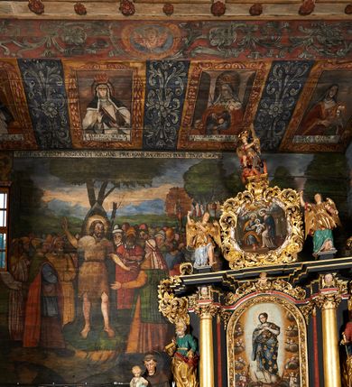 Zdjęcie nr 48: Polichromia ścienna prezbiterium pokrywa wszystkie ściany oraz strop tej części świątyni. 

Polichromia ścienna stropu
Strop zdobiony jest malowanymi kasetonami, w których umieszczone są rozety na różnokolorowych tłach. W centrum ukazane są dwie prostokątne kompozycje, ujęte w ornamentalne ramy. Od wschodu znajduje się Zwiastowanie Zachariaszowi, powyżej inskrypcja: „NE TIMEAS ZACHARIA VXOR TVA PARIET TIBI / FILIVM ET VOCABIS NOMEN EIVS IOANNEM LUCAE 1”. W drugiej kompozycji przedstawiono Nawiedzenie z Najświętszej Marii Panny ze św. Zachariaszem i św. Józefem, powyżej inskrypcja: „EXVLTAVIT IN GAVDIO INFANS IN VTERO MEO LUCAE 1”. 

Polichromia ścienna ścian prezbiterium
Dekoracje ścian o formie pasowej. W dolnej kondygnacji malowane iluzjonistycznie portrety świętych węgierskich w ornamentalnych ramach. Postacie ukazane do wysokości kolan, zwrócone trzy czwarte z odpowiednimi im atrybutami w dłoniach. Portrety opatrzone u góry łacińskimi opisami. Na ścianie północnej galerię otwiera wizerunek króla Stefana z napisem: „S(ANCTVS) STEPHANVS REX VNGARIAE, REGNVM SVVM DEIPARAE / DONAVIT OBIIT. 1038. 20 AVG(VSTVS) SEP. ALBERE 9.”. Następnie ukazani są kolejno: św. Emeryk „S(ANCTVS) EMERIGVS DVX VNGARIAE VT VIRGO MANERET, / A. CAELO PERSVASVS OBIIT. 1032. 5 nou. Sen. Albdvewas”, św. Władysław: „S(ANCTVS) LADISLAVS REX UNGARIAE ROSARIO ENSI OBVO/LUTO PVGNARE SOLITVS OBIIT 1097. 27.  iun. Sep. varad.”. Po drugiej stronie prezbiterium umieszczone są cztery obrazy świętych o tych samych wymiarach, również opatrzone napisami. Od lewej strony kolejno: św. Salomon „S(ANCTVS) SALOMON REX VNGARIAE DEIN EREMITA VITAE / AVSTERITATE FLORVIT OBIIT 1077. 28. Sept. Sep. Pold. in Idria”, św. Kazimierz: „S(ANCTVS) CASIMIRVS ELECTVS REX VNGARIAE AMORE / SARVANDAE VIRGINITATIS OBIIT 1484 4. MARTII.”, św. Dawid: „S(ANTCVS) DAVID REX SCOTIAE, FILIVS S(ANCTA) MARGA/RITAE VNGARA MATRE NATAE OBIIT 1153”, św. Ludwik: „S(SANCTVS) LVDOVICVS TOLOSANVS EP. E MARIA / REGIS VNG. FILIA NATVS OBIIT 1297 19 Aug(ustvs)”. Cykl ten kończy przy ścianie tęczowej przedstawienie św. Gintera „S(ANCVS) GVNTHERVS EREM. S(ANCTVS) STEPHANI REGIS / VNGARIAE COGNATVS OBIIT 1045 9. oct.”. Dodatkowo w obrazy wpisane są zacheuszki w formie równoramiennych krzyży ujętych w medaliony utworzone z wieńca. W zamknięciu prezbiterium przedstawienia diabłów, a od zachodu pod amboną czyściec.

Kondygnacja druga o podziałach architektonicznych u dołu wydzielona cokołem, dekorowanym złotymi girlandami na czarnym tle. Prostokątne pola wydzielone za pomocą malowanych iluzjonistycznie kolumn korynckich dźwigających architraw zdobiony stylizowanym kimationem jońskim. Powyżej fryz wypełniony wicią akantową, pomiędzy którą wplecione są kartusze z przedstawieniami świętych. W polach, pomiędzy kolumnami wielofigurowe kompozycje ze scenami z życia św. Jana Chrzciciela: Głos wołającego na pustyni „TV QVIS ES VOX CLAMANTIS IN DESERTO IOANN(IS) 1.”, Oto Baranek Boży „ECCE AGNVS DEI ECCE QVI TOLLIT PECCATVM MVNDI IOANN. IMÕ.”, Chrzest Jezusa w Jordanie „IESVS BAPTISTAVS EST A IONNE IN IORDANE MARCI. 1.”, Św. Jan Chrzciciel napomina Heroda „DICEBAT IOANNES HERODI NON LICET TIBI HABERE VXOREM FRATRIS TVI. MARCI 6,19.”, Św. Maria Magdalena pokutująca na pustyni, Św. Jan Chrzciciel na pustyni, Św. Jan Chrzciciel wtrącony do więzienia „HERODES TENVIT IONNEM ET VINXIT EVM IN CARCERE MAR 67o”, Kat podaje głowę św. Jana Chrzciciela Salome „DECOLLAVIT EVM IN CARCERE ET DEDIT CAPVT PVELLAE MAR.6.(?)”, Uczta Heroda „HERODES NATALIS SVI COENAM FECIT PRINCIPIBVS MAR. 6 10.”, Salome podaje głowę św. Jana Chrzciciela matce „CAPVT EIVS PVELLA DEDIT MATRI SVE M. 61”. 

W ostatniej, trzeciej strefie, umieszczonej w partii fryzu znajdują się medaliony. Na ścianie tęczowej, następnie na ścianie północnej i południowej ukazani są kolejno: św. Maciej „SA[N]CTVS / MATTHIAS”, Chrystus „IESVS / CHRISTVS”, św. Piotr „SANCTVS / PETRVS”, św. Szymon „SANCTVS / SIMON”, św. Andrzej „SANCTVS / ANDREAS”, św. Jakub Większy „S(ANCTVS) IACOBI / MAIORI”, św. Jan „(?)”, św. Filip „(?)”, św. Bartłomiej „SANCTVS / BARTHOLO/MAVS”, św. Tomasz „SANCTVS / THOMAS”, św. Mateusz „SANCTVS / MATTHAEVS”, św. Jakub Mniejszy „S(ANCTVS) IACOBVS / MINOR” i św. Juda Tadeusz „S(ANCVS) IVDA / THADEVS” na ścianie tęczowej od południa. Na belce tęczowej, od strony prezbiterium w wieńcu laurowym napis: „PICTVM / A[NNO] D[OMINI] 1711”.