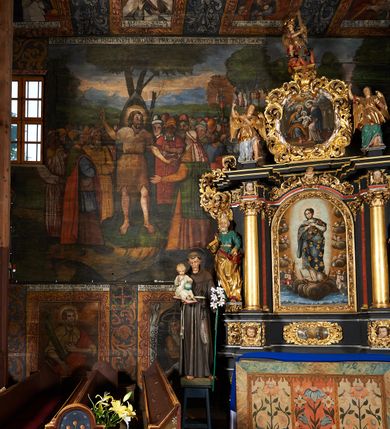 Zdjęcie nr 49: Polichromia ścienna prezbiterium pokrywa wszystkie ściany oraz strop tej części świątyni. 

Polichromia ścienna stropu
Strop zdobiony jest malowanymi kasetonami, w których umieszczone są rozety na różnokolorowych tłach. W centrum ukazane są dwie prostokątne kompozycje, ujęte w ornamentalne ramy. Od wschodu znajduje się Zwiastowanie Zachariaszowi, powyżej inskrypcja: „NE TIMEAS ZACHARIA VXOR TVA PARIET TIBI / FILIVM ET VOCABIS NOMEN EIVS IOANNEM LUCAE 1”. W drugiej kompozycji przedstawiono Nawiedzenie z Najświętszej Marii Panny ze św. Zachariaszem i św. Józefem, powyżej inskrypcja: „EXVLTAVIT IN GAVDIO INFANS IN VTERO MEO LUCAE 1”. 

Polichromia ścienna ścian prezbiterium
Dekoracje ścian o formie pasowej. W dolnej kondygnacji malowane iluzjonistycznie portrety świętych węgierskich w ornamentalnych ramach. Postacie ukazane do wysokości kolan, zwrócone trzy czwarte z odpowiednimi im atrybutami w dłoniach. Portrety opatrzone u góry łacińskimi opisami. Na ścianie północnej galerię otwiera wizerunek króla Stefana z napisem: „S(ANCTVS) STEPHANVS REX VNGARIAE, REGNVM SVVM DEIPARAE / DONAVIT OBIIT. 1038. 20 AVG(VSTVS) SEP. ALBERE 9.”. Następnie ukazani są kolejno: św. Emeryk „S(ANCTVS) EMERIGVS DVX VNGARIAE VT VIRGO MANERET, / A. CAELO PERSVASVS OBIIT. 1032. 5 nou. Sen. Albdvewas”, św. Władysław: „S(ANCTVS) LADISLAVS REX UNGARIAE ROSARIO ENSI OBVO/LUTO PVGNARE SOLITVS OBIIT 1097. 27.  iun. Sep. varad.”. Po drugiej stronie prezbiterium umieszczone są cztery obrazy świętych o tych samych wymiarach, również opatrzone napisami. Od lewej strony kolejno: św. Salomon „S(ANCTVS) SALOMON REX VNGARIAE DEIN EREMITA VITAE / AVSTERITATE FLORVIT OBIIT 1077. 28. Sept. Sep. Pold. in Idria”, św. Kazimierz: „S(ANCTVS) CASIMIRVS ELECTVS REX VNGARIAE AMORE / SARVANDAE VIRGINITATIS OBIIT 1484 4. MARTII.”, św. Dawid: „S(ANTCVS) DAVID REX SCOTIAE, FILIVS S(ANCTA) MARGA/RITAE VNGARA MATRE NATAE OBIIT 1153”, św. Ludwik: „S(SANCTVS) LVDOVICVS TOLOSANVS EP. E MARIA / REGIS VNG. FILIA NATVS OBIIT 1297 19 Aug(ustvs)”. Cykl ten kończy przy ścianie tęczowej przedstawienie św. Gintera „S(ANCVS) GVNTHERVS EREM. S(ANCTVS) STEPHANI REGIS / VNGARIAE COGNATVS OBIIT 1045 9. oct.”. Dodatkowo w obrazy wpisane są zacheuszki w formie równoramiennych krzyży ujętych w medaliony utworzone z wieńca. W zamknięciu prezbiterium przedstawienia diabłów, a od zachodu pod amboną czyściec.

Kondygnacja druga o podziałach architektonicznych u dołu wydzielona cokołem, dekorowanym złotymi girlandami na czarnym tle. Prostokątne pola wydzielone za pomocą malowanych iluzjonistycznie kolumn korynckich dźwigających architraw zdobiony stylizowanym kimationem jońskim. Powyżej fryz wypełniony wicią akantową, pomiędzy którą wplecione są kartusze z przedstawieniami świętych. W polach, pomiędzy kolumnami wielofigurowe kompozycje ze scenami z życia św. Jana Chrzciciela: Głos wołającego na pustyni „TV QVIS ES VOX CLAMANTIS IN DESERTO IOANN(IS) 1.”, Oto Baranek Boży „ECCE AGNVS DEI ECCE QVI TOLLIT PECCATVM MVNDI IOANN. IMÕ.”, Chrzest Jezusa w Jordanie „IESVS BAPTISTAVS EST A IONNE IN IORDANE MARCI. 1.”, Św. Jan Chrzciciel napomina Heroda „DICEBAT IOANNES HERODI NON LICET TIBI HABERE VXOREM FRATRIS TVI. MARCI 6,19.”, Św. Maria Magdalena pokutująca na pustyni, Św. Jan Chrzciciel na pustyni, Św. Jan Chrzciciel wtrącony do więzienia „HERODES TENVIT IONNEM ET VINXIT EVM IN CARCERE MAR 67o”, Kat podaje głowę św. Jana Chrzciciela Salome „DECOLLAVIT EVM IN CARCERE ET DEDIT CAPVT PVELLAE MAR.6.(?)”, Uczta Heroda „HERODES NATALIS SVI COENAM FECIT PRINCIPIBVS MAR. 6 10.”, Salome podaje głowę św. Jana Chrzciciela matce „CAPVT EIVS PVELLA DEDIT MATRI SVE M. 61”. 

W ostatniej, trzeciej strefie, umieszczonej w partii fryzu znajdują się medaliony. Na ścianie tęczowej, następnie na ścianie północnej i południowej ukazani są kolejno: św. Maciej „SA[N]CTVS / MATTHIAS”, Chrystus „IESVS / CHRISTVS”, św. Piotr „SANCTVS / PETRVS”, św. Szymon „SANCTVS / SIMON”, św. Andrzej „SANCTVS / ANDREAS”, św. Jakub Większy „S(ANCTVS) IACOBI / MAIORI”, św. Jan „(?)”, św. Filip „(?)”, św. Bartłomiej „SANCTVS / BARTHOLO/MAVS”, św. Tomasz „SANCTVS / THOMAS”, św. Mateusz „SANCTVS / MATTHAEVS”, św. Jakub Mniejszy „S(ANCTVS) IACOBVS / MINOR” i św. Juda Tadeusz „S(ANCVS) IVDA / THADEVS” na ścianie tęczowej od południa. Na belce tęczowej, od strony prezbiterium w wieńcu laurowym napis: „PICTVM / A[NNO] D[OMINI] 1711”.