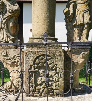 Zdjęcie nr 4: Kapliczka ustawiona na niskim prostopadłościennym postumencie. Pośrodku na wysokim cokole umieszczona kolumna, na której szczycie ustawiona jest figura Matki Boskiej Niepokalanie Poczętej z Dzieciątkiem. Cokół flankowany szerokimi spływami wolutowymi, na których ustawione są na niskich podstawach rzeźby: po lewej św. Donata, a po prawej św. Floriana. Na przedniej ściance cokołu znajduje się płaskorzeźba św. Marii Magdaleny ujęta płyciną w kształcie stojącego prostokąta, zamkniętego łukiem nadwieszonym z uskokami. Święta ukazana jest na tle groty, zwrócona trzy czwarte w prawo z rozpuszczonymi włosami, z dyscypliną w dłoni. Po lewej stronie kompozycji znajduje się wysoki, prosty krzyż i czaszka. Nad płyciną napis „ANNO D(OMI)NI 1758 / S(ANCTA) MARIA MAGDALENA / Zda SPPIPM”. Na tylnej ściance cokołu rzeźba św. Rozalii, ukazana w podobnym ujęciu z napisem „SANCTA ROSALIA VIRGO”. Święty Donat ukazany w postawie stojącej w delikatnym kontrapoście z księgą w lewej dłoni i trójzębem w prawej. Ubrany jest w strój biskupi: albę, komżę, kapę, infułę, rękawice i buty. Ma szeroką twarz, okoloną krótką brodą, bujne i długie włosy spływające na plecy. Na podstawie napis: „S(ANCTUS) DONATUS”. Po prawej stronie kolumny rzeźba św. Floriana, ustawionego frontalnie, który prawą ręką polewa wodą płonący dom, a lewą podtrzymuje u ramienia połę płaszcza. Ubrany jest w strój legionisty rzymskiego z hełmem o wyraźnie podkreślonym pióropuszu. Twarz szeroka, okolona średniej długości włosami, schowanymi pod hełmem. Podstawa pod rzeźbą opleciona metalowymi ściskami. Kolumna ustawiona na bazie, trzon gładki, dekorowany w połowie wysokości dwiema uskrzydlonymi główkami aniołów na tle promienistej glorii, kapitel koryncki. Rzeźba Matki Boskiej Niepokalanej z Dzieciątkiem na prawym ręku ustawiona jest frontalnie, w delikatnym kontrapoście na globie ziemskim, oplecionym przez węża. Maria ubrana jest w długą suknię i silnie drapowany płaszcz. Twarz owalna, okolona bujnymi i długimi włosami. Dzieciątko zwrócone jest w stronę Matki Boskiej. Rzeźba Marii osłonięta jest metalowym daszkiem wspartym na czterech prętach przytwierdzonych do metalowej opaski oplatającej kapitel kolumny. Kapliczka obwiedziona metalowym płotkiem. 

