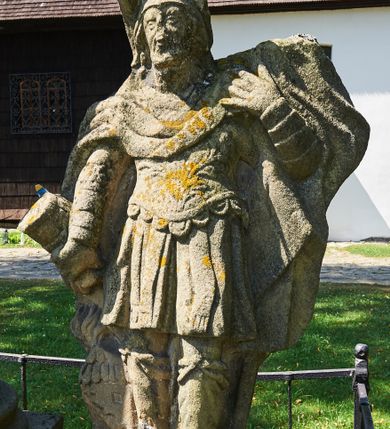 Zdjęcie nr 5: Kapliczka ustawiona na niskim prostopadłościennym postumencie. Pośrodku na wysokim cokole umieszczona kolumna, na której szczycie ustawiona jest figura Matki Boskiej Niepokalanie Poczętej z Dzieciątkiem. Cokół flankowany szerokimi spływami wolutowymi, na których ustawione są na niskich podstawach rzeźby: po lewej św. Donata, a po prawej św. Floriana. Na przedniej ściance cokołu znajduje się płaskorzeźba św. Marii Magdaleny ujęta płyciną w kształcie stojącego prostokąta, zamkniętego łukiem nadwieszonym z uskokami. Święta ukazana jest na tle groty, zwrócona trzy czwarte w prawo z rozpuszczonymi włosami, z dyscypliną w dłoni. Po lewej stronie kompozycji znajduje się wysoki, prosty krzyż i czaszka. Nad płyciną napis „ANNO D(OMI)NI 1758 / S(ANCTA) MARIA MAGDALENA / Zda SPPIPM”. Na tylnej ściance cokołu rzeźba św. Rozalii, ukazana w podobnym ujęciu z napisem „SANCTA ROSALIA VIRGO”. Święty Donat ukazany w postawie stojącej w delikatnym kontrapoście z księgą w lewej dłoni i trójzębem w prawej. Ubrany jest w strój biskupi: albę, komżę, kapę, infułę, rękawice i buty. Ma szeroką twarz, okoloną krótką brodą, bujne i długie włosy spływające na plecy. Na podstawie napis: „S(ANCTUS) DONATUS”. Po prawej stronie kolumny rzeźba św. Floriana, ustawionego frontalnie, który prawą ręką polewa wodą płonący dom, a lewą podtrzymuje u ramienia połę płaszcza. Ubrany jest w strój legionisty rzymskiego z hełmem o wyraźnie podkreślonym pióropuszu. Twarz szeroka, okolona średniej długości włosami, schowanymi pod hełmem. Podstawa pod rzeźbą opleciona metalowymi ściskami. Kolumna ustawiona na bazie, trzon gładki, dekorowany w połowie wysokości dwiema uskrzydlonymi główkami aniołów na tle promienistej glorii, kapitel koryncki. Rzeźba Matki Boskiej Niepokalanej z Dzieciątkiem na prawym ręku ustawiona jest frontalnie, w delikatnym kontrapoście na globie ziemskim, oplecionym przez węża. Maria ubrana jest w długą suknię i silnie drapowany płaszcz. Twarz owalna, okolona bujnymi i długimi włosami. Dzieciątko zwrócone jest w stronę Matki Boskiej. Rzeźba Marii osłonięta jest metalowym daszkiem wspartym na czterech prętach przytwierdzonych do metalowej opaski oplatającej kapitel kolumny. Kapliczka obwiedziona metalowym płotkiem. 

