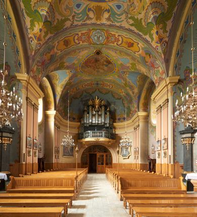 Zdjęcie nr 4: Chór muzyczny przyścienny, podwieszony na ścianie chóru zachodniego, wsparty na dwóch wklęsło-wypukłych konsolach z motywami palmet i kartuszem pośrodku. Parapet chóru wybrzuszony w części środkowej, z wklęsłymi bokami; od spodu aplikowana przestylizowana, stiukowa dekoracja roślinna. Parapet z tralkami powyżej, poprzedzielanymi sześcioma filarkami; gzyms prowadzony analogicznie jak podstawa chóru. Prospekt organowy pięcioosiowy, jednokondygnacyjny ze zwieńczeniem, z osiami skrajnymi i środkową równej wysokości oraz osiami drugą i czwartą wyższymi od pozostałych. Cokół prospektu wcięty na bokach, na osiach skrajnych zbiegający ku środkowi, w trzech osiach środkowych w kształcie półkolumienki. Uszy w formie delikatnych spływów z piszczałkami wewnątrz; piszczałki w każdej osi ustawione podobnie jak cokół prospektu. Belkowanie niepełne, wyłamane; gzyms w osiach bocznych zbiegający ku środkowi, w trzech osiach środkowych lekko wybrzuszony. Zwieńczenia kwater z piszczałkami dekorowane imitacją kratki regencyjnej, podobnie jak pola z piszczałkami pomiędzy osiami oraz zwieńczenia każdej z pięciu osi. Nad osią środkową wielka gloria promienista z motywem maski solarnej pośrodku.

Podstawa chóru malowana na odcień ciemnej żółci, parapet z tralkami w odcieniach ciemnej szarości, podobnie jak prospekt. Detale złocone.