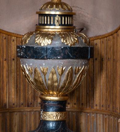 Zdjęcie nr 1: Chrzcielnica w typie kielichowym osadzona jest na stopie na planie koła. Walcowaty, krótki trzon ujęty jest pierścieniem i zakończony wałkiem. Czarę w formie spłaszczonej od góry kuli objęto cęgą. W dolnej części dekorowana jest naprzemiennie płaskorzeźbionymi, smukłymi liśćmi i kwiatami na długich łodygach, zaś w górnej rozłożystymi liśćmi stylizowanego akantu. Pokrywa jest w formie baldachimu, z kopułką na walcowatej podstawie, dekorowanego u dołu pasem kanelur, u góry spłaszczonymi puklowaniami i zwieńczona szyszką z krzyżem. Całość jest marmoryzowana, w kolorze ciemnoszarym i szaroróżowym, detale i ornamentyka złocona.