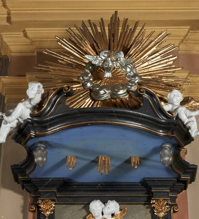 Zdjęcie nr 8: Ambona przyścienna, wisząca z dostępem od strony wewnętrznej klatki schodowej usytuowanej w murze kościoła. Kosz na planie kwadratu o wklęsłych bokach, wypukłym froncie i ściętych narożach, z podwieszeniem w formie stylizowanego, odwróconego stożka zakończonego wolutą. Kosz w narożach ujęty pilastrami o trzonach w formie wolut, dźwigającymi belkowanie parapetu. Ścianki dekorowane płycinami z polami o obrysie utworzonym z linii wklęsło-wypukłej wzbogaconej odcinkami prostymi; w płycinach uskrzydlone główki anielskie. W partii belkowania parapetu front ambony zdobiony muszlą, ujętą kwiatowymi girlandami, na tle pilastrów dwa putta. Zaplecek ujęty pilastrami oraz spływami wolutowymi, dźwigającymi belkowanie. W polu drzwiczki w formie stojącego prostokąta, dekorowane płyciną o wykroju ukształtowanym na bazie falistej, wklęsło-wypukłej linii. Powyżej dwie uskrzydlone główki anielskie. Baldachim płaski, na planie analogicznym do kosza ambony, opasany profilowanym gzymsem, dźwigającym przerwany, wolutowy przyczółek, flankowany przez dwa putta. Całość zwieńczona glorią promienistą z rzeźbioną gołębicą Ducha Świętego.

 Struktura marmoryzowana, w kolorze brązowo-szarym, ciemnoszarym i niebieskim, profile i ornamenty złocone i srebrzone.