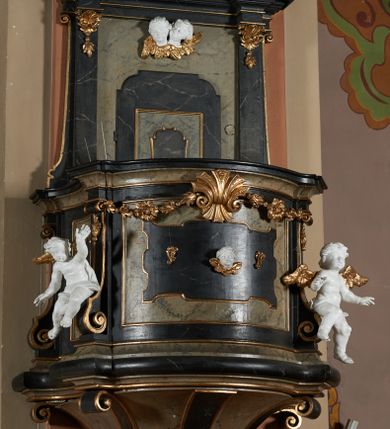 Zdjęcie nr 1: Ambona przyścienna, wisząca z dostępem od strony wewnętrznej klatki schodowej usytuowanej w murze kościoła. Kosz na planie kwadratu o wklęsłych bokach, wypukłym froncie i ściętych narożach, z podwieszeniem w formie stylizowanego, odwróconego stożka zakończonego wolutą. Kosz w narożach ujęty pilastrami o trzonach w formie wolut, dźwigającymi belkowanie parapetu. Ścianki dekorowane płycinami z polami o obrysie utworzonym z linii wklęsło-wypukłej wzbogaconej odcinkami prostymi; w płycinach uskrzydlone główki anielskie. W partii belkowania parapetu front ambony zdobiony muszlą, ujętą kwiatowymi girlandami, na tle pilastrów dwa putta. Zaplecek ujęty pilastrami oraz spływami wolutowymi, dźwigającymi belkowanie. W polu drzwiczki w formie stojącego prostokąta, dekorowane płyciną o wykroju ukształtowanym na bazie falistej, wklęsło-wypukłej linii. Powyżej dwie uskrzydlone główki anielskie. Baldachim płaski, na planie analogicznym do kosza ambony, opasany profilowanym gzymsem, dźwigającym przerwany, wolutowy przyczółek, flankowany przez dwa putta. Całość zwieńczona glorią promienistą z rzeźbioną gołębicą Ducha Świętego.

 Struktura marmoryzowana, w kolorze brązowo-szarym, ciemnoszarym i niebieskim, profile i ornamenty złocone i srebrzone.