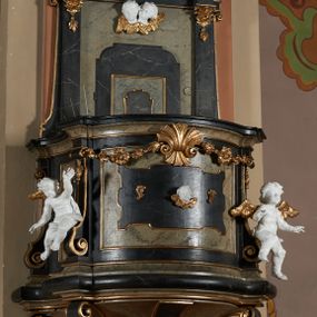 Zdjęcie nr 1: Ambona przyścienna, wisząca z dostępem od strony wewnętrznej klatki schodowej usytuowanej w murze kościoła. Kosz na planie kwadratu o wklęsłych bokach, wypukłym froncie i ściętych narożach, z podwieszeniem w formie stylizowanego, odwróconego stożka zakończonego wolutą. Kosz w narożach ujęty pilastrami o trzonach w formie wolut, dźwigającymi belkowanie parapetu. Ścianki dekorowane płycinami z polami o obrysie utworzonym z linii wklęsło-wypukłej wzbogaconej odcinkami prostymi; w płycinach uskrzydlone główki anielskie. W partii belkowania parapetu front ambony zdobiony muszlą, ujętą kwiatowymi girlandami, na tle pilastrów dwa putta. Zaplecek ujęty pilastrami oraz spływami wolutowymi, dźwigającymi belkowanie. W polu drzwiczki w formie stojącego prostokąta, dekorowane płyciną o wykroju ukształtowanym na bazie falistej, wklęsło-wypukłej linii. Powyżej dwie uskrzydlone główki anielskie. Baldachim płaski, na planie analogicznym do kosza ambony, opasany profilowanym gzymsem, dźwigającym przerwany, wolutowy przyczółek, flankowany przez dwa putta. Całość zwieńczona glorią promienistą z rzeźbioną gołębicą Ducha Świętego.

 Struktura marmoryzowana, w kolorze brązowo-szarym, ciemnoszarym i niebieskim, profile i ornamenty złocone i srebrzone.