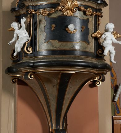 Zdjęcie nr 5: Ambona przyścienna, wisząca z dostępem od strony wewnętrznej klatki schodowej usytuowanej w murze kościoła. Kosz na planie kwadratu o wklęsłych bokach, wypukłym froncie i ściętych narożach, z podwieszeniem w formie stylizowanego, odwróconego stożka zakończonego wolutą. Kosz w narożach ujęty pilastrami o trzonach w formie wolut, dźwigającymi belkowanie parapetu. Ścianki dekorowane płycinami z polami o obrysie utworzonym z linii wklęsło-wypukłej wzbogaconej odcinkami prostymi; w płycinach uskrzydlone główki anielskie. W partii belkowania parapetu front ambony zdobiony muszlą, ujętą kwiatowymi girlandami, na tle pilastrów dwa putta. Zaplecek ujęty pilastrami oraz spływami wolutowymi, dźwigającymi belkowanie. W polu drzwiczki w formie stojącego prostokąta, dekorowane płyciną o wykroju ukształtowanym na bazie falistej, wklęsło-wypukłej linii. Powyżej dwie uskrzydlone główki anielskie. Baldachim płaski, na planie analogicznym do kosza ambony, opasany profilowanym gzymsem, dźwigającym przerwany, wolutowy przyczółek, flankowany przez dwa putta. Całość zwieńczona glorią promienistą z rzeźbioną gołębicą Ducha Świętego.

 Struktura marmoryzowana, w kolorze brązowo-szarym, ciemnoszarym i niebieskim, profile i ornamenty złocone i srebrzone.