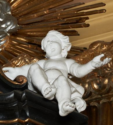 Zdjęcie nr 11: Ambona przyścienna, wisząca z dostępem od strony wewnętrznej klatki schodowej usytuowanej w murze kościoła. Kosz na planie kwadratu o wklęsłych bokach, wypukłym froncie i ściętych narożach, z podwieszeniem w formie stylizowanego, odwróconego stożka zakończonego wolutą. Kosz w narożach ujęty pilastrami o trzonach w formie wolut, dźwigającymi belkowanie parapetu. Ścianki dekorowane płycinami z polami o obrysie utworzonym z linii wklęsło-wypukłej wzbogaconej odcinkami prostymi; w płycinach uskrzydlone główki anielskie. W partii belkowania parapetu front ambony zdobiony muszlą, ujętą kwiatowymi girlandami, na tle pilastrów dwa putta. Zaplecek ujęty pilastrami oraz spływami wolutowymi, dźwigającymi belkowanie. W polu drzwiczki w formie stojącego prostokąta, dekorowane płyciną o wykroju ukształtowanym na bazie falistej, wklęsło-wypukłej linii. Powyżej dwie uskrzydlone główki anielskie. Baldachim płaski, na planie analogicznym do kosza ambony, opasany profilowanym gzymsem, dźwigającym przerwany, wolutowy przyczółek, flankowany przez dwa putta. Całość zwieńczona glorią promienistą z rzeźbioną gołębicą Ducha Świętego.

 Struktura marmoryzowana, w kolorze brązowo-szarym, ciemnoszarym i niebieskim, profile i ornamenty złocone i srebrzone.