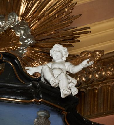 Zdjęcie nr 10: Ambona przyścienna, wisząca z dostępem od strony wewnętrznej klatki schodowej usytuowanej w murze kościoła. Kosz na planie kwadratu o wklęsłych bokach, wypukłym froncie i ściętych narożach, z podwieszeniem w formie stylizowanego, odwróconego stożka zakończonego wolutą. Kosz w narożach ujęty pilastrami o trzonach w formie wolut, dźwigającymi belkowanie parapetu. Ścianki dekorowane płycinami z polami o obrysie utworzonym z linii wklęsło-wypukłej wzbogaconej odcinkami prostymi; w płycinach uskrzydlone główki anielskie. W partii belkowania parapetu front ambony zdobiony muszlą, ujętą kwiatowymi girlandami, na tle pilastrów dwa putta. Zaplecek ujęty pilastrami oraz spływami wolutowymi, dźwigającymi belkowanie. W polu drzwiczki w formie stojącego prostokąta, dekorowane płyciną o wykroju ukształtowanym na bazie falistej, wklęsło-wypukłej linii. Powyżej dwie uskrzydlone główki anielskie. Baldachim płaski, na planie analogicznym do kosza ambony, opasany profilowanym gzymsem, dźwigającym przerwany, wolutowy przyczółek, flankowany przez dwa putta. Całość zwieńczona glorią promienistą z rzeźbioną gołębicą Ducha Świętego.

 Struktura marmoryzowana, w kolorze brązowo-szarym, ciemnoszarym i niebieskim, profile i ornamenty złocone i srebrzone.