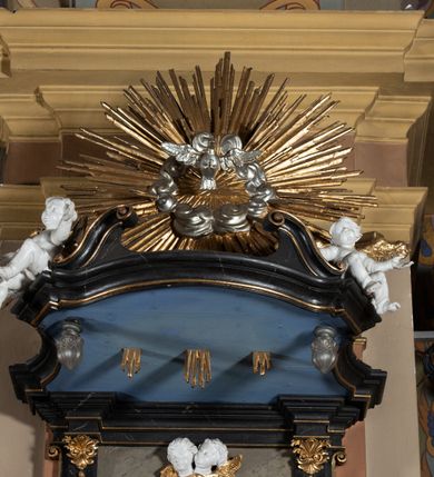 Zdjęcie nr 7: Ambona przyścienna, wisząca z dostępem od strony wewnętrznej klatki schodowej usytuowanej w murze kościoła. Kosz na planie kwadratu o wklęsłych bokach, wypukłym froncie i ściętych narożach, z podwieszeniem w formie stylizowanego, odwróconego stożka zakończonego wolutą. Kosz w narożach ujęty pilastrami o trzonach w formie wolut, dźwigającymi belkowanie parapetu. Ścianki dekorowane płycinami z polami o obrysie utworzonym z linii wklęsło-wypukłej wzbogaconej odcinkami prostymi; w płycinach uskrzydlone główki anielskie. W partii belkowania parapetu front ambony zdobiony muszlą, ujętą kwiatowymi girlandami, na tle pilastrów dwa putta. Zaplecek ujęty pilastrami oraz spływami wolutowymi, dźwigającymi belkowanie. W polu drzwiczki w formie stojącego prostokąta, dekorowane płyciną o wykroju ukształtowanym na bazie falistej, wklęsło-wypukłej linii. Powyżej dwie uskrzydlone główki anielskie. Baldachim płaski, na planie analogicznym do kosza ambony, opasany profilowanym gzymsem, dźwigającym przerwany, wolutowy przyczółek, flankowany przez dwa putta. Całość zwieńczona glorią promienistą z rzeźbioną gołębicą Ducha Świętego.

 Struktura marmoryzowana, w kolorze brązowo-szarym, ciemnoszarym i niebieskim, profile i ornamenty złocone i srebrzone.