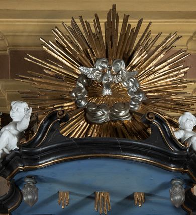 Zdjęcie nr 9: Ambona przyścienna, wisząca z dostępem od strony wewnętrznej klatki schodowej usytuowanej w murze kościoła. Kosz na planie kwadratu o wklęsłych bokach, wypukłym froncie i ściętych narożach, z podwieszeniem w formie stylizowanego, odwróconego stożka zakończonego wolutą. Kosz w narożach ujęty pilastrami o trzonach w formie wolut, dźwigającymi belkowanie parapetu. Ścianki dekorowane płycinami z polami o obrysie utworzonym z linii wklęsło-wypukłej wzbogaconej odcinkami prostymi; w płycinach uskrzydlone główki anielskie. W partii belkowania parapetu front ambony zdobiony muszlą, ujętą kwiatowymi girlandami, na tle pilastrów dwa putta. Zaplecek ujęty pilastrami oraz spływami wolutowymi, dźwigającymi belkowanie. W polu drzwiczki w formie stojącego prostokąta, dekorowane płyciną o wykroju ukształtowanym na bazie falistej, wklęsło-wypukłej linii. Powyżej dwie uskrzydlone główki anielskie. Baldachim płaski, na planie analogicznym do kosza ambony, opasany profilowanym gzymsem, dźwigającym przerwany, wolutowy przyczółek, flankowany przez dwa putta. Całość zwieńczona glorią promienistą z rzeźbioną gołębicą Ducha Świętego.

 Struktura marmoryzowana, w kolorze brązowo-szarym, ciemnoszarym i niebieskim, profile i ornamenty złocone i srebrzone.