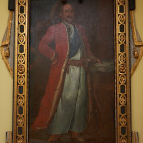 Zdjęcie nr 1: Obraz w kształcie stojącego prostokąta ujętego dekoracyjną ramą z uszakami, motywem paludamentum oraz wyobrażeniem Orderu Orła Białego przedstawia całopostaciową sylwetkę Franciszka Szwracenberg-Czernego. Twarz, zwróconego w trzech czwartych w prawo mężczyzny charakteryzują pozbawione idealizacji cechy portretowe o długim, zaokrąglonym na końcu nosie, lekko podkrążonych oczach, szerokich ustach, spiczastych wąsach, bruzdach na czole oraz wydatnym podbródku. Ubrany jest on w długi, biały żupan przewiązany czerwonym pasem kontuszowym oraz czerwoną, podszytą futrem gronostajowym delię. Przez piersi przewieszona jest błękitna szarfa będąca fragmentem garnituru Orderu Orła Białego. U lewego boku przypięta jest szabla. Prawą rękę wspiera na biodrze, lewą, w której trzyma rysunek fasady kościoła, na niewielkim stoliku o wygiętych nogach. W tle, po lewej stronie kompozycji widoczna jest granatowa, obszyta złotą frędzlą kotara.     