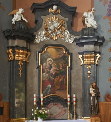 Zdjęcie nr 1: Nastawa ołtarza stiukowa, architektoniczna, jednoosiowa, jednokondygnacyjna, o wklęsłym licu, ze zwieńczeniem oraz osadzona na wysokim cokole. Ołtarz drewniany, sarkofagowy z drewnianą, silnie wysuniętą mensą. Retabulum ujęte parą zdwojonych pilastrów o trzonach dekorowanych konsolkami i kampanulami na froncie, dźwigających przerwane belkowanie. W polu środkowym umieszczono obraz Świętej Rodziny ujęty profilowaną ramą w formie stojącego prostokąta zamkniętego łukiem odcinkowym, nadwieszonym z uskokiem i uszakami po bokach, wklęsłego u spodu. Poniżej obrazu znajduje się płycina ujęta spływami wolutowymi, zamknięta fragmentem gzymsu w formie łuku odcinkowego. W jej polu jest kartusz o fantazyjnym wykroju. Zwieńczenie ujęto parą spływów wolutowych i zamknięto fragmentem wklęsło-wypukłego gzymsu, na którym jest płomienisty wazon. Na końcach spływów wolutowych znajduje się para siedzących putt.  
Pole zwieńczenia wypełniają płaskorzeźbione obłoki z uskrzydlonymi główkami anielskimi oraz ukazana na ich tle promienista gloria z parą główek anielskich pośrodku. Struktura retabulum jest marmoryzowana w kolorze szaro-brązowym i ciemnoszarym, ze złoconymi i srebrzonymi detalami.
