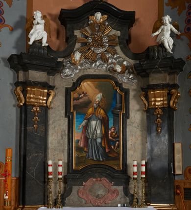 Zdjęcie nr 1: Nastawa ołtarza stiukowa, architektoniczna, jednoosiowa, jednokondygnacyjna, o wklęsłym licu, ze zwieńczeniem oraz osadzona na wysokim cokole. Ołtarz drewniany, sarkofagowy z drewnianą, silnie wysuniętą mensą. Retabulum ujęte jest parą zdwojonych pilastrów o trzonach dekorowanych konsolkami i kampanulami na froncie, dźwigających przerwane belkowanie. W polu środkowym umieszczono obraz św. Mikołaja ujęty profilowaną ramą w formie stojącego prostokąta zamkniętego łukiem odcinkowym, nadwieszonym z uskokiem i uszakami po bokach, wklęsłego u spodu. Poniżej obrazu znajduje się płycina ujęta spływami wolutowymi, zamknięta fragmentem gzymsu w formie łuku odcinkowego. W jej polu jest kartusz o fantazyjnym wykroju. Zwieńczenie ujęto parą spływów wolutowych i zamknięto fragmentem wklęsło-wypukłego gzymsu, na którym jest płomienisty wazon. Na końcach spływów wolutowych znajduje się para siedzących putt.  
Pole zwieńczenia wypełniają płaskorzeźbione obłoki z uskrzydlonymi główkami anielskimi oraz ukazana na ich tle promienista gloria z monogramem maryjnym pośrodku. Struktura retabulum jest marmoryzowana w kolorze szaro-brązowym i ciemnoszarym, ze złoconymi i srebrzonymi detalami.
