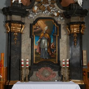 Zdjęcie nr 1: Nastawa ołtarza stiukowa, architektoniczna, jednoosiowa, jednokondygnacyjna, o wklęsłym licu, ze zwieńczeniem oraz osadzona na wysokim cokole. Ołtarz drewniany, sarkofagowy z drewnianą, silnie wysuniętą mensą. Retabulum ujęte jest parą zdwojonych pilastrów o trzonach dekorowanych konsolkami i kampanulami na froncie, dźwigających przerwane belkowanie. W polu środkowym umieszczono obraz św. Mikołaja ujęty profilowaną ramą w formie stojącego prostokąta zamkniętego łukiem odcinkowym, nadwieszonym z uskokiem i uszakami po bokach, wklęsłego u spodu. Poniżej obrazu znajduje się płycina ujęta spływami wolutowymi, zamknięta fragmentem gzymsu w formie łuku odcinkowego. W jej polu jest kartusz o fantazyjnym wykroju. Zwieńczenie ujęto parą spływów wolutowych i zamknięto fragmentem wklęsło-wypukłego gzymsu, na którym jest płomienisty wazon. Na końcach spływów wolutowych znajduje się para siedzących putt.  
Pole zwieńczenia wypełniają płaskorzeźbione obłoki z uskrzydlonymi główkami anielskimi oraz ukazana na ich tle promienista gloria z monogramem maryjnym pośrodku. Struktura retabulum jest marmoryzowana w kolorze szaro-brązowym i ciemnoszarym, ze złoconymi i srebrzonymi detalami.
