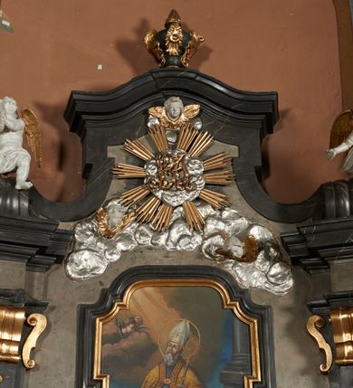 Zdjęcie nr 28: Nastawa ołtarza stiukowa, architektoniczna, jednoosiowa, jednokondygnacyjna, o wklęsłym licu, ze zwieńczeniem oraz osadzona na wysokim cokole. Ołtarz drewniany, sarkofagowy z drewnianą, silnie wysuniętą mensą. Retabulum ujęte jest parą zdwojonych pilastrów o trzonach dekorowanych konsolkami i kampanulami na froncie, dźwigających przerwane belkowanie. W polu środkowym umieszczono obraz św. Mikołaja ujęty profilowaną ramą w formie stojącego prostokąta zamkniętego łukiem odcinkowym, nadwieszonym z uskokiem i uszakami po bokach, wklęsłego u spodu. Poniżej obrazu znajduje się płycina ujęta spływami wolutowymi, zamknięta fragmentem gzymsu w formie łuku odcinkowego. W jej polu jest kartusz o fantazyjnym wykroju. Zwieńczenie ujęto parą spływów wolutowych i zamknięto fragmentem wklęsło-wypukłego gzymsu, na którym jest płomienisty wazon. Na końcach spływów wolutowych znajduje się para siedzących putt.  
Pole zwieńczenia wypełniają płaskorzeźbione obłoki z uskrzydlonymi główkami anielskimi oraz ukazana na ich tle promienista gloria z monogramem maryjnym pośrodku. Struktura retabulum jest marmoryzowana w kolorze szaro-brązowym i ciemnoszarym, ze złoconymi i srebrzonymi detalami.

