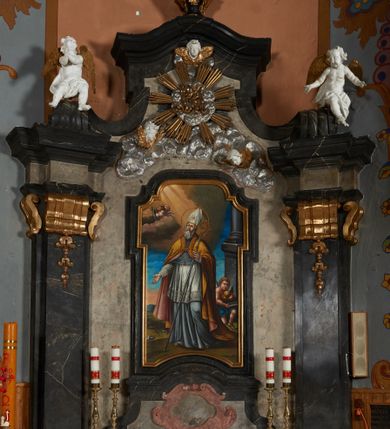 Zdjęcie nr 13: Nastawa ołtarza stiukowa, architektoniczna, jednoosiowa, jednokondygnacyjna, o wklęsłym licu, ze zwieńczeniem oraz osadzona na wysokim cokole. Ołtarz drewniany, sarkofagowy z drewnianą, silnie wysuniętą mensą. Retabulum ujęte jest parą zdwojonych pilastrów o trzonach dekorowanych konsolkami i kampanulami na froncie, dźwigających przerwane belkowanie. W polu środkowym umieszczono obraz św. Mikołaja ujęty profilowaną ramą w formie stojącego prostokąta zamkniętego łukiem odcinkowym, nadwieszonym z uskokiem i uszakami po bokach, wklęsłego u spodu. Poniżej obrazu znajduje się płycina ujęta spływami wolutowymi, zamknięta fragmentem gzymsu w formie łuku odcinkowego. W jej polu jest kartusz o fantazyjnym wykroju. Zwieńczenie ujęto parą spływów wolutowych i zamknięto fragmentem wklęsło-wypukłego gzymsu, na którym jest płomienisty wazon. Na końcach spływów wolutowych znajduje się para siedzących putt.  
Pole zwieńczenia wypełniają płaskorzeźbione obłoki z uskrzydlonymi główkami anielskimi oraz ukazana na ich tle promienista gloria z monogramem maryjnym pośrodku. Struktura retabulum jest marmoryzowana w kolorze szaro-brązowym i ciemnoszarym, ze złoconymi i srebrzonymi detalami.
