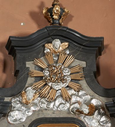 Zdjęcie nr 24: Nastawa ołtarza stiukowa, architektoniczna, jednoosiowa, jednokondygnacyjna, o wklęsłym licu, ze zwieńczeniem oraz osadzona na wysokim cokole. Ołtarz drewniany, sarkofagowy z drewnianą, silnie wysuniętą mensą. Retabulum ujęte jest parą zdwojonych pilastrów o trzonach dekorowanych konsolkami i kampanulami na froncie, dźwigających przerwane belkowanie. W polu środkowym umieszczono obraz św. Mikołaja ujęty profilowaną ramą w formie stojącego prostokąta zamkniętego łukiem odcinkowym, nadwieszonym z uskokiem i uszakami po bokach, wklęsłego u spodu. Poniżej obrazu znajduje się płycina ujęta spływami wolutowymi, zamknięta fragmentem gzymsu w formie łuku odcinkowego. W jej polu jest kartusz o fantazyjnym wykroju. Zwieńczenie ujęto parą spływów wolutowych i zamknięto fragmentem wklęsło-wypukłego gzymsu, na którym jest płomienisty wazon. Na końcach spływów wolutowych znajduje się para siedzących putt.  
Pole zwieńczenia wypełniają płaskorzeźbione obłoki z uskrzydlonymi główkami anielskimi oraz ukazana na ich tle promienista gloria z monogramem maryjnym pośrodku. Struktura retabulum jest marmoryzowana w kolorze szaro-brązowym i ciemnoszarym, ze złoconymi i srebrzonymi detalami.
