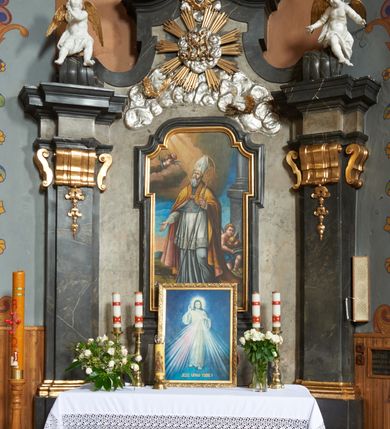Zdjęcie nr 2: Nastawa ołtarza stiukowa, architektoniczna, jednoosiowa, jednokondygnacyjna, o wklęsłym licu, ze zwieńczeniem oraz osadzona na wysokim cokole. Ołtarz drewniany, sarkofagowy z drewnianą, silnie wysuniętą mensą. Retabulum ujęte jest parą zdwojonych pilastrów o trzonach dekorowanych konsolkami i kampanulami na froncie, dźwigających przerwane belkowanie. W polu środkowym umieszczono obraz św. Mikołaja ujęty profilowaną ramą w formie stojącego prostokąta zamkniętego łukiem odcinkowym, nadwieszonym z uskokiem i uszakami po bokach, wklęsłego u spodu. Poniżej obrazu znajduje się płycina ujęta spływami wolutowymi, zamknięta fragmentem gzymsu w formie łuku odcinkowego. W jej polu jest kartusz o fantazyjnym wykroju. Zwieńczenie ujęto parą spływów wolutowych i zamknięto fragmentem wklęsło-wypukłego gzymsu, na którym jest płomienisty wazon. Na końcach spływów wolutowych znajduje się para siedzących putt.  
Pole zwieńczenia wypełniają płaskorzeźbione obłoki z uskrzydlonymi główkami anielskimi oraz ukazana na ich tle promienista gloria z monogramem maryjnym pośrodku. Struktura retabulum jest marmoryzowana w kolorze szaro-brązowym i ciemnoszarym, ze złoconymi i srebrzonymi detalami.
