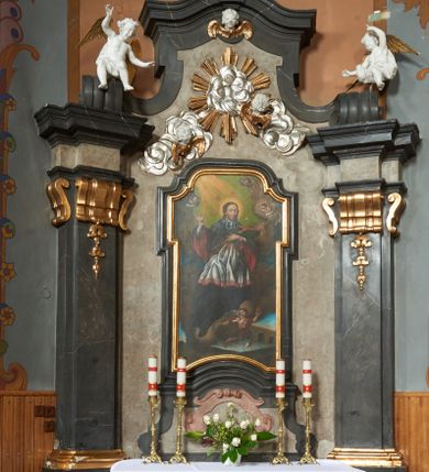 Zdjęcie nr 1: Nastawa ołtarza stiukowa, architektoniczna, jednoosiowa, jednokondygnacyjna, o wklęsłym licu, ze zwieńczeniem oraz osadzona na wysokim cokole. Ołtarz drewniany, sarkofagowy z drewnianą, silnie wysuniętą mensą. Retabulum ujęte parą zdwojonych pilastrów o trzonach dekorowanych konsolkami i kampanulami na froncie, dźwigających przerwane belkowanie. W polu środkowym umieszczono obraz św. Jana Nepomucena ujęty profilowaną ramą w formie stojącego prostokąta zamkniętego łukiem odcinkowym, nadwieszonym z uskokiem i uszakami po bokach, wklęsłego u spodu. Poniżej obrazu znajduje się płycina ujęta spływami wolutowymi, zamknięta fragmentem gzymsu w formie łuku odcinkowego. W jej polu jest kartusz o fantazyjnym wykroju. Zwieńczenie ujęto parą spływów wolutowych i zamknięto fragmentem wklęsło-wypukłego gzymsu, na którym jest krzyż na kuli. Na końcach spływów wolutowych znajduje się para siedzących putt.  
Pole zwieńczenia wypełniają płaskorzeźbione obłoki z uskrzydlonymi główkami anielskimi oraz ukazana na ich tle promienista gloria. Struktura retabulum jest marmoryzowana w kolorze szaro-brązowym i ciemnoszarym, ze złoconymi i srebrzonymi detalami.

