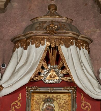 Zdjęcie nr 11: Architektoniczna nastawa ołtarzowa jest jednoosiowa, jednokondygnacyjna ze zwieńczeniem, osadzona na cokole, ujęta parą kolumn o kapitelach kompozytowych, wysuniętych przed strukturę. Każda usytuowana jest na tle pary wklęsłych, ustawionych pod kątem pilastrów, zdwojonych po zewnętrznej stronie nastawy. Kolumny i pilastry wspierają przerwane belkowanie. Pole środkowe retabulum zaaranżowane jest w formie paludamentu pod baldachimem, którego krawędź ozdobiono lambrekinem. Tkanina po bokach jest podwiązana wstęgami. Na tle paludamentu znajduje się niewielka wnęka w kształcie stojącego prostokąta, ujęta profilowaną, dekorowaną ornamentem ramą z wizerunkiem Matki Boskiej z Dzieciątkiem. Ponad obrazem umieszczono płaskorzeźbione oko Opatrzności Bożej ukazane na tle obłoków i glorii promienistej. Zwieńczenie ołtarza ujęte jest parą wolut spływających na belkowanie i zamknięte fragmentem gzymsu w formie łuku odcinkowego zakończonego wolutami. Po bokach znajduje się para adorujących aniołów. W polu zwieńczenia zlokalizowano owalny medalion z monogramem maryjnym, ujęty promienistą glorią oraz otokiem z obłoków i uskrzydlonych główek anielskich. Nastawę flankują cokoły z figurami św. Anny i św. Joachima. Struktura retabulum jest marmoryzowana w kolorze szaro-brązowym i ciemnoróżowym, detale polichromowane na biało, złocone i srebrzone.