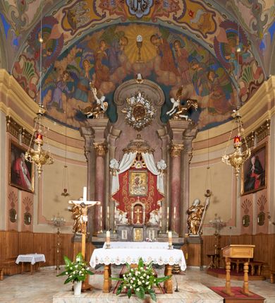 Zdjęcie nr 2: Architektoniczna nastawa ołtarzowa jest jednoosiowa, jednokondygnacyjna ze zwieńczeniem, osadzona na cokole, ujęta parą kolumn o kapitelach kompozytowych, wysuniętych przed strukturę. Każda usytuowana jest na tle pary wklęsłych, ustawionych pod kątem pilastrów, zdwojonych po zewnętrznej stronie nastawy. Kolumny i pilastry wspierają przerwane belkowanie. Pole środkowe retabulum zaaranżowane jest w formie paludamentu pod baldachimem, którego krawędź ozdobiono lambrekinem. Tkanina po bokach jest podwiązana wstęgami. Na tle paludamentu znajduje się niewielka wnęka w kształcie stojącego prostokąta, ujęta profilowaną, dekorowaną ornamentem ramą z wizerunkiem Matki Boskiej z Dzieciątkiem. Ponad obrazem umieszczono płaskorzeźbione oko Opatrzności Bożej ukazane na tle obłoków i glorii promienistej. Zwieńczenie ołtarza ujęte jest parą wolut spływających na belkowanie i zamknięte fragmentem gzymsu w formie łuku odcinkowego zakończonego wolutami. Po bokach znajduje się para adorujących aniołów. W polu zwieńczenia zlokalizowano owalny medalion z monogramem maryjnym, ujęty promienistą glorią oraz otokiem z obłoków i uskrzydlonych główek anielskich. Nastawę flankują cokoły z figurami św. Anny i św. Joachima. Struktura retabulum jest marmoryzowana w kolorze szaro-brązowym i ciemnoróżowym, detale polichromowane na biało, złocone i srebrzone.