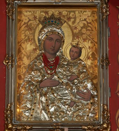 Zdjęcie nr 2: Obraz w kształcie stojącego prostokąta przedstawia Matkę Boską z Dzieciątkiem wyobrażoną w półpostaci. Sylwetka Marii nieznacznie zwrócona jest w lewo. Jej zaokrągloną twarz charakteryzują łagodne, wyraziste rysy o silnie podkreślonych liniach łuków brwiowych i prostego nosa wydobyte światłocieniem. Oczy są duże, usta drobne, a karnacja śniada. Partie szat, łącznie z maforionem zasłaniającym włosy Marii, pokryte są metalowymi, srebrzonymi sukienkami, na powierzchni których odtworzono w ogólnych zarysach przebieg fałdów; szaty zdobione są motywem stylizowanej, ukwieconej, złoconej wici roślinnej, o kameryzowanych lamówkach. Maria prawą ręką o wyprostowanych palcach wskazuje na trzymane na lewej ręce Dzieciątko, w trzech czwartych skierowane w lewo. Zwróconą niemal na wprost twarz charakteryzuje podobny jak w przypadku Marii sposób modelunku, okalają ją krótkie, brązowe włosy. Ubraną w długą tunikę sylwetkę Jezusa także osłania metalowa sukienka. Lewą ręką podtrzymuje on na kolanach księgę, prawą unosi w geście błogosławieństwa; u dołu widoczna jest nieobuta, swobodnie zwisająca stopa Dzieciątka. Postaci ukazane są na tłoczonym w gruncie, złoconym tle dekorowanym motywem roślinnym. Zwieńczone otwartymi, kameryzowanymi koronami głowy okalają nimby i wieńce z gwiazd. Wizerunek ujęty profilowaną, srebrzoną ramą zdobioną złoconymi motywami rocaille.