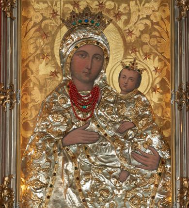 Zdjęcie nr 1: Obraz w kształcie stojącego prostokąta przedstawia Matkę Boską z Dzieciątkiem wyobrażoną w półpostaci. Sylwetka Marii nieznacznie zwrócona jest w lewo. Jej zaokrągloną twarz charakteryzują łagodne, wyraziste rysy o silnie podkreślonych liniach łuków brwiowych i prostego nosa wydobyte światłocieniem. Oczy są duże, usta drobne, a karnacja śniada. Partie szat, łącznie z maforionem zasłaniającym włosy Marii, pokryte są metalowymi, srebrzonymi sukienkami, na powierzchni których odtworzono w ogólnych zarysach przebieg fałdów; szaty zdobione są motywem stylizowanej, ukwieconej, złoconej wici roślinnej, o kameryzowanych lamówkach. Maria prawą ręką o wyprostowanych palcach wskazuje na trzymane na lewej ręce Dzieciątko, w trzech czwartych skierowane w lewo. Zwróconą niemal na wprost twarz charakteryzuje podobny jak w przypadku Marii sposób modelunku, okalają ją krótkie, brązowe włosy. Ubraną w długą tunikę sylwetkę Jezusa także osłania metalowa sukienka. Lewą ręką podtrzymuje on na kolanach księgę, prawą unosi w geście błogosławieństwa; u dołu widoczna jest nieobuta, swobodnie zwisająca stopa Dzieciątka. Postaci ukazane są na tłoczonym w gruncie, złoconym tle dekorowanym motywem roślinnym. Zwieńczone otwartymi, kameryzowanymi koronami głowy okalają nimby i wieńce z gwiazd. Wizerunek ujęty profilowaną, srebrzoną ramą zdobioną złoconymi motywami rocaille.