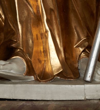 Zdjęcie nr 6: Figura ustawiona na na kwadratowej podstawie, drążona z tyłu, przedstawia stojącego św. Joachima, wyobrażonego w całej postaci. zwróconego nieznacznie w prawo, z uniesioną ku górze głową. Prawa ręka św. Joachima spoczywa na piersi, w lewej, lekko zgiętej w łokciu i odsuniętej w bok trzyma szufle pasterską. Twarz jest pociągła, wychudzona, z długim zarostem i o wyrazistych rysach z silnie uwypuklonymi kośćmi policzkowymi, liniami łuków brwiowych, wąskim nosem, małymi, rozchylonymi, pełnymi ustami z uniesioną górną wargą oraz dużymi, głęboko osadzonymi oczami spoglądającymi w górę. Krótkie, lekko falowane włosy układają się w regularne pukle. Smukłą i wydłużoną sylwetkę świętego osłania przylegająca do ciała, długa, drapowana suknia przepasana w talii oraz narzucony na plecy i opadający wzdłuż boków płaszcz z fragmentem tkaniny zasłaniającym częściowo lewe udo. Kolorystyka odsłoniętych partii ciała utrzymana jest w bieli, szaty złocone, drzewiec, na którym osadzona jest szufla srebrzony.