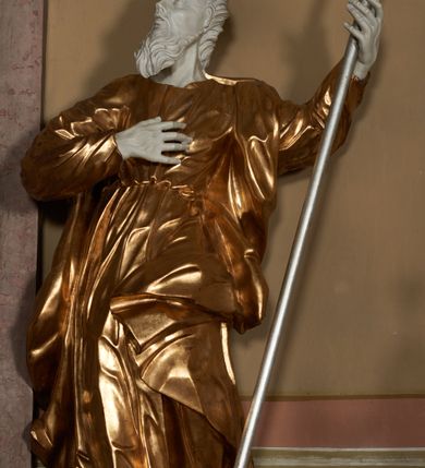 Zdjęcie nr 2: Figura ustawiona na na kwadratowej podstawie, drążona z tyłu, przedstawia stojącego św. Joachima, wyobrażonego w całej postaci. zwróconego nieznacznie w prawo, z uniesioną ku górze głową. Prawa ręka św. Joachima spoczywa na piersi, w lewej, lekko zgiętej w łokciu i odsuniętej w bok trzyma szufle pasterską. Twarz jest pociągła, wychudzona, z długim zarostem i o wyrazistych rysach z silnie uwypuklonymi kośćmi policzkowymi, liniami łuków brwiowych, wąskim nosem, małymi, rozchylonymi, pełnymi ustami z uniesioną górną wargą oraz dużymi, głęboko osadzonymi oczami spoglądającymi w górę. Krótkie, lekko falowane włosy układają się w regularne pukle. Smukłą i wydłużoną sylwetkę świętego osłania przylegająca do ciała, długa, drapowana suknia przepasana w talii oraz narzucony na plecy i opadający wzdłuż boków płaszcz z fragmentem tkaniny zasłaniającym częściowo lewe udo. Kolorystyka odsłoniętych partii ciała utrzymana jest w bieli, szaty złocone, drzewiec, na którym osadzona jest szufla srebrzony.