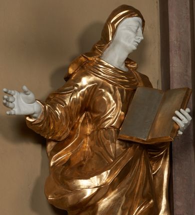 Zdjęcie nr 1: Figura ustawiona wprost na kwadratowej podstawie, drążona z tyłu, przedstawia św. Annę, wyobrażoną w całej postaci, stojącą w kontrapoście, zwróconą nieznacznie w prawo, z głową pochyloną i skierowaną w lewo. Św. Anna lewą ręką podtrzymuje wspartą na piersi, otwartą księgę, prawa lekko zgięta w łokciu wyciągnięta jest w bok. Twarz o wyrazistych, dynamiczne modelowanych rysach oraz o szczupłej fizjonomii podkreślona jest uwydatnionymi kośćmi policzkowymi, liniami łuków brwiowych, długim, wąskim nosem, wąskimi, rozchylonymi ustami z lekko uniesioną górną wargą oraz dużymi, głęboko osadzonymi oczami. Sylwetka jest wydłużona i smukła. Święta ubrana jest w ściśle przylegającą do ciała, obficie drapowaną suknię z szerokim, fałdowanym kołnierzem, na lewe ramię narzucony ma osłaniający sylwetkę płaszcz. Na głowę nałożony jest rozwiany welon. Odsłonięte partie ciała utrzymane są w bieli, szaty i atrybuty złocone.