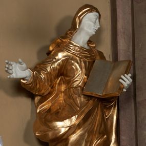 Zdjęcie nr 1: Figura ustawiona wprost na kwadratowej podstawie, drążona z tyłu, przedstawia św. Annę, wyobrażoną w całej postaci, stojącą w kontrapoście, zwróconą nieznacznie w prawo, z głową pochyloną i skierowaną w lewo. Św. Anna lewą ręką podtrzymuje wspartą na piersi, otwartą księgę, prawa lekko zgięta w łokciu wyciągnięta jest w bok. Twarz o wyrazistych, dynamiczne modelowanych rysach oraz o szczupłej fizjonomii podkreślona jest uwydatnionymi kośćmi policzkowymi, liniami łuków brwiowych, długim, wąskim nosem, wąskimi, rozchylonymi ustami z lekko uniesioną górną wargą oraz dużymi, głęboko osadzonymi oczami. Sylwetka jest wydłużona i smukła. Święta ubrana jest w ściśle przylegającą do ciała, obficie drapowaną suknię z szerokim, fałdowanym kołnierzem, na lewe ramię narzucony ma osłaniający sylwetkę płaszcz. Na głowę nałożony jest rozwiany welon. Odsłonięte partie ciała utrzymane są w bieli, szaty i atrybuty złocone.