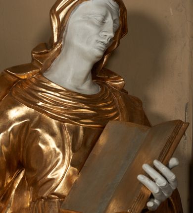 Zdjęcie nr 3: Figura ustawiona wprost na kwadratowej podstawie, drążona z tyłu, przedstawia św. Annę, wyobrażoną w całej postaci, stojącą w kontrapoście, zwróconą nieznacznie w prawo, z głową pochyloną i skierowaną w lewo. Św. Anna lewą ręką podtrzymuje wspartą na piersi, otwartą księgę, prawa lekko zgięta w łokciu wyciągnięta jest w bok. Twarz o wyrazistych, dynamiczne modelowanych rysach oraz o szczupłej fizjonomii podkreślona jest uwydatnionymi kośćmi policzkowymi, liniami łuków brwiowych, długim, wąskim nosem, wąskimi, rozchylonymi ustami z lekko uniesioną górną wargą oraz dużymi, głęboko osadzonymi oczami. Sylwetka jest wydłużona i smukła. Święta ubrana jest w ściśle przylegającą do ciała, obficie drapowaną suknię z szerokim, fałdowanym kołnierzem, na lewe ramię narzucony ma osłaniający sylwetkę płaszcz. Na głowę nałożony jest rozwiany welon. Odsłonięte partie ciała utrzymane są w bieli, szaty i atrybuty złocone.