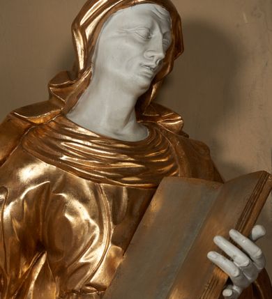 Zdjęcie nr 4: Figura ustawiona wprost na kwadratowej podstawie, drążona z tyłu, przedstawia św. Annę, wyobrażoną w całej postaci, stojącą w kontrapoście, zwróconą nieznacznie w prawo, z głową pochyloną i skierowaną w lewo. Św. Anna lewą ręką podtrzymuje wspartą na piersi, otwartą księgę, prawa lekko zgięta w łokciu wyciągnięta jest w bok. Twarz o wyrazistych, dynamiczne modelowanych rysach oraz o szczupłej fizjonomii podkreślona jest uwydatnionymi kośćmi policzkowymi, liniami łuków brwiowych, długim, wąskim nosem, wąskimi, rozchylonymi ustami z lekko uniesioną górną wargą oraz dużymi, głęboko osadzonymi oczami. Sylwetka jest wydłużona i smukła. Święta ubrana jest w ściśle przylegającą do ciała, obficie drapowaną suknię z szerokim, fałdowanym kołnierzem, na lewe ramię narzucony ma osłaniający sylwetkę płaszcz. Na głowę nałożony jest rozwiany welon. Odsłonięte partie ciała utrzymane są w bieli, szaty i atrybuty złocone.