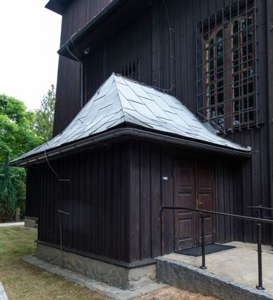 Zdjęcie nr 6: Kościół drewniany, orientowany, jednonawowy o konstrukcji zrębowej, oszalowany. Prezbiterium kwadratowe o wielobocznym zamknięciu z zakrystią od północy. Nawa szersza, prawie kwadratowa, przy niej od północy kaplica i składzik później dostawiony; od południa i od zachodu kruchty. Korpus nakryty dachem dwuspadowym, jednokalenicowym, z barokową wieżyczką na sygnaturkę na kalenicy; składzik nakryty dachem pulpitowym. Wewnątrz stropy kasetonowe. Otwór tęczy o profilowanym łuku półkolistym podwyższonym; belka profilowana, na niej grupa Ukrzyżowania. Chór muzyczny wsparty na dwóch słupach. Kaplica otwarta do nawy półkolistą arkadą. Troje drzwi o ozdobnych obramieniach uszatych, schodkowatych i uszato-schodkowatych, ze starymi okuciami. Wieża nowa, izbicowa o pochyłych ścianach, zwieńczona ostrosłupowym hełmem.
