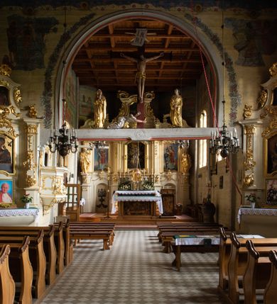 Zdjęcie nr 26: Kościół drewniany, orientowany, jednonawowy o konstrukcji zrębowej, oszalowany. Prezbiterium kwadratowe o wielobocznym zamknięciu z zakrystią od północy. Nawa szersza, prawie kwadratowa, przy niej od północy kaplica i składzik później dostawiony; od południa i od zachodu kruchty. Korpus nakryty dachem dwuspadowym, jednokalenicowym, z barokową wieżyczką na sygnaturkę na kalenicy; składzik nakryty dachem pulpitowym. Wewnątrz stropy kasetonowe. Otwór tęczy o profilowanym łuku półkolistym podwyższonym; belka profilowana, na niej grupa Ukrzyżowania. Chór muzyczny wsparty na dwóch słupach. Kaplica otwarta do nawy półkolistą arkadą. Troje drzwi o ozdobnych obramieniach uszatych, schodkowatych i uszato-schodkowatych, ze starymi okuciami. Wieża nowa, izbicowa o pochyłych ścianach, zwieńczona ostrosłupowym hełmem.
