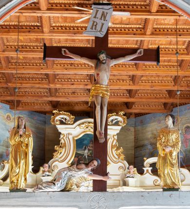 Zdjęcie nr 1: Łuk tęczowy o półkolistym wykroju, na profilowanej belce zdobionej pośrodku ryzowaną rozetą ustawiona grupa Ukrzyżowania. W centrum krucyfiks, po prawej św. Jan Ewangelista, po lewej Matka Boska i św. Maria Magdalena obejmująca krzyż. Chrystus przybity do krzyża trzema gwoździami, sylwetka w delikatnym zwisie, z głową pochyloną delikatnie w dół, o sztywnym układzie sylwetki z zaznaczonymi śladami męki. Perizonium w formie tkaniny przewiązanej na prawym biodrze ze zwisem wzdłuż prawego uda. Twarz podłużna, okolona zarostem; włosy długie spływające na prawe ramię w silnie skręcone pukle, na głowie zielona korona cierniowa złożona z dwóch przeplatających się gałązek. Nad głową Chrystusa biały titulus z napisem: „I(ESUS) N(AZARENUS) R(EX) I(UDAEORUM)”. Krzyż prosty, gładki o ramionach zakończonych gzymsikami. Po lewej stronie krucyfiksu rzeźba św. Marii Magdaleny ukazana w pozycji klęczącej, pochylonej do przodu, obejmująca dłońmi krzyż. Święta jest ubrana w długą, srebrzoną suknię, odsłaniającą nagie ramiona, przewiązaną w talii. Twarz szeroka, delikatnie otwarte usta, małe oczy i długi nos. Głowę okalają długie, ciemne włosy, spływające na plecy. Za nią ustawiona jest rzeźba Matki Boskiej w delikatnym kontrapoście, z prawą nogą ugiętą w kolanie, ze splecionymi na wysokości piersi rękami. Maria jest ubrana w długą, złoconą suknię oraz złocony płaszcz założony na głowę i spływający wzdłuż sylwetki. Twarz szeroka, z wyraźnie zaznaczonym podbródkiem, wzrok skierowany na krucyfiks, zmarszczone brwi. Po prawej stronie rzeźba św. Jana Ewangelisty, ukazanego w postawie stojącej, zwróconego w trzech czwartych w prawo, z rękami splecionymi na wysokości pasa. Twarz owalna, małe usta, długi i wąski nos, wzrok skierowany ku górze, zmarszczone brwi. Włosy ciemne, bujne, spływające na plecy. Święty ubrany jest w długą, złoconą suknię z długimi rękawami. Rzeźby ustawione są na niskich, zielonych cokolikach. Polichromia w odsłoniętych partiach ciała naturalistyczna, detale złocone i srebrzone. 
