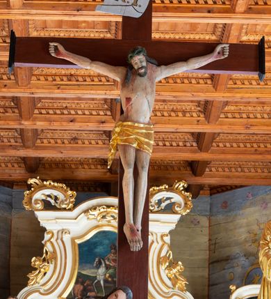 Zdjęcie nr 2: Łuk tęczowy o półkolistym wykroju, na profilowanej belce zdobionej pośrodku ryzowaną rozetą ustawiona grupa Ukrzyżowania. W centrum krucyfiks, po prawej św. Jan Ewangelista, po lewej Matka Boska i św. Maria Magdalena obejmująca krzyż. Chrystus przybity do krzyża trzema gwoździami, sylwetka w delikatnym zwisie, z głową pochyloną delikatnie w dół, o sztywnym układzie sylwetki z zaznaczonymi śladami męki. Perizonium w formie tkaniny przewiązanej na prawym biodrze ze zwisem wzdłuż prawego uda. Twarz podłużna, okolona zarostem; włosy długie spływające na prawe ramię w silnie skręcone pukle, na głowie zielona korona cierniowa złożona z dwóch przeplatających się gałązek. Nad głową Chrystusa biały titulus z napisem: „I(ESUS) N(AZARENUS) R(EX) I(UDAEORUM)”. Krzyż prosty, gładki o ramionach zakończonych gzymsikami. Po lewej stronie krucyfiksu rzeźba św. Marii Magdaleny ukazana w pozycji klęczącej, pochylonej do przodu, obejmująca dłońmi krzyż. Święta jest ubrana w długą, srebrzoną suknię, odsłaniającą nagie ramiona, przewiązaną w talii. Twarz szeroka, delikatnie otwarte usta, małe oczy i długi nos. Głowę okalają długie, ciemne włosy, spływające na plecy. Za nią ustawiona jest rzeźba Matki Boskiej w delikatnym kontrapoście, z prawą nogą ugiętą w kolanie, ze splecionymi na wysokości piersi rękami. Maria jest ubrana w długą, złoconą suknię oraz złocony płaszcz założony na głowę i spływający wzdłuż sylwetki. Twarz szeroka, z wyraźnie zaznaczonym podbródkiem, wzrok skierowany na krucyfiks, zmarszczone brwi. Po prawej stronie rzeźba św. Jana Ewangelisty, ukazanego w postawie stojącej, zwróconego w trzech czwartych w prawo, z rękami splecionymi na wysokości pasa. Twarz owalna, małe usta, długi i wąski nos, wzrok skierowany ku górze, zmarszczone brwi. Włosy ciemne, bujne, spływające na plecy. Święty ubrany jest w długą, złoconą suknię z długimi rękawami. Rzeźby ustawione są na niskich, zielonych cokolikach. Polichromia w odsłoniętych partiach ciała naturalistyczna, detale złocone i srebrzone. 
